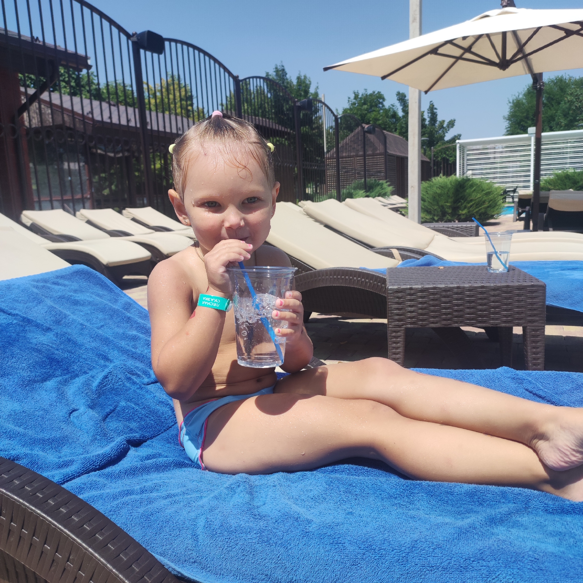 A young girl with blonde hair styled in pigtails is sitting on a bright blue towel on a lounge chair by a pool. She is wearing a light blue swimsuit with pink trim. She is holding a clear plastic cup with a blue straw and drinking from it. Water droplets are visible on her skin. A teal wristband is on her left wrist with Russian text that appears to say "ПЕСЧАНАЯ СКАЗКА" (Peschanaya Skazka), which translates to "Sandy Fairy Tale." In the background, there are rows of empty lounge chairs, a dark brown woven table with a glass of water and a straw, a black metal fence, lush green trees, and a large beige umbrella providing shade. The sky is clear and blue, suggesting a sunny day. The scene appears to be a resort or a water park. The girl's expression is somewhat playful.