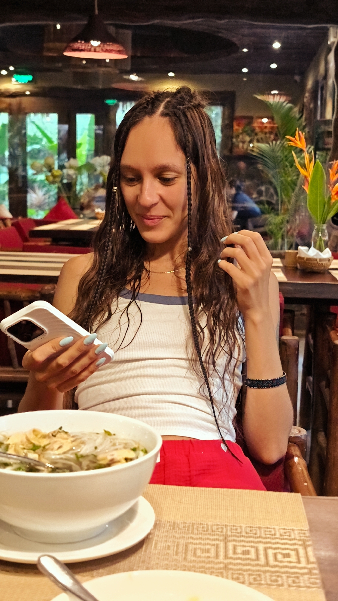A young woman is seated at a table in what appears to be a restaurant. She is holding a white smartphone in her left hand and looking down at it. Her nails are painted a light blue. She is wearing a white tank top with a blue trim and red pants. Her hair is styled in braids. In front of her on the table is a bowl of soup and a placemat. The background shows a restaurant interior with wooden furniture, plants, and lighting fixtures. The location is identified as Nam Hoa Lư Ward, Vietnam.