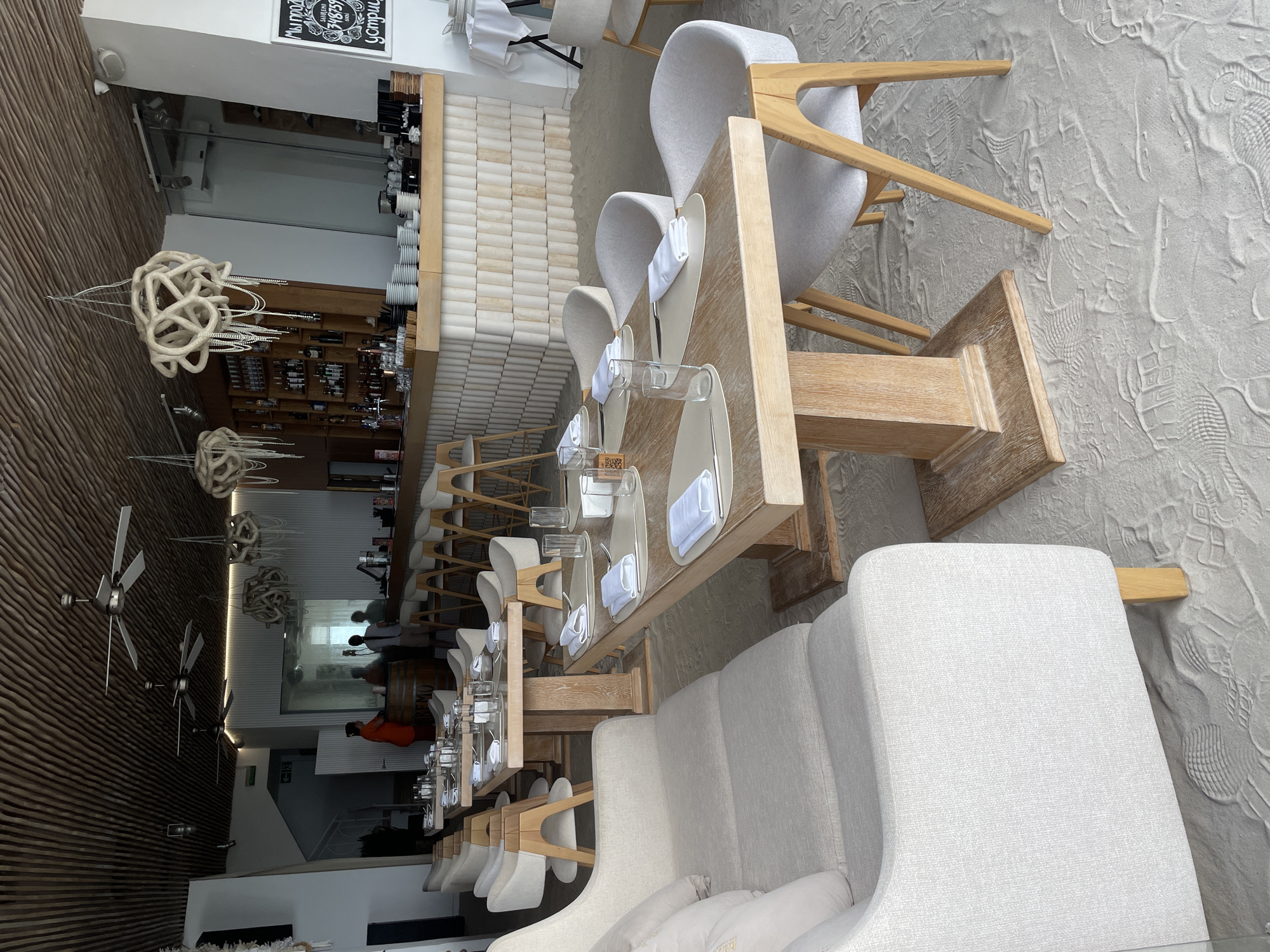 The image shows the interior of a modern restaurant with a minimalist aesthetic. The scene is brightly lit, suggesting it is daytime, though no windows are clearly visible to confirm natural light. The restaurant appears to be set up for dining, with several tables and chairs arranged. One long table is set with plates, white napkins, and glasses, indicating that patrons are either being served or have recently dined.
Objects in the scene include light-colored upholstered chairs and sofas, a wooden table, and decorative hanging light fixtures that resemble tangled rope. In the background, a bar area is visible with stools and bottles displayed on shelves. The floor has a textured, sandy appearance, possibly an intentional design choice to evoke a beach or natural setting. There are also ceiling fans mounted on a wood-paneled wall.
A few people are present in the restaurant. Two individuals are visible in the background near the bar, appearing to be staff members or possibly patrons. Another person is seen standing in a more dimly lit area towards the back, wearing an orange shirt. The overall atmosphere seems calm and sophisticated.
A small sign with text is visible on the wall near the top left of the image, but the text is blurry and illegible. The location is identified as Sochi, Russia.