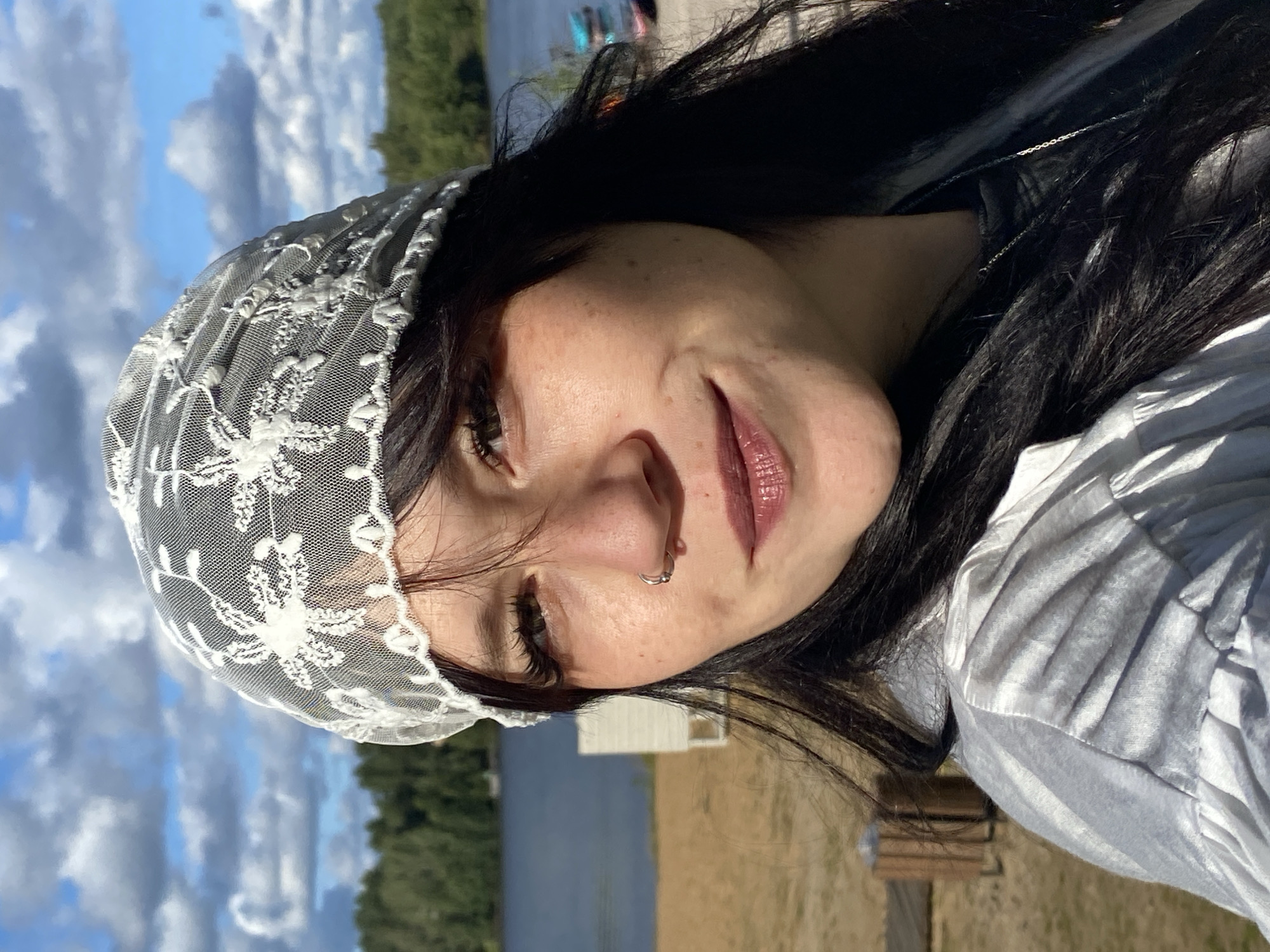 A medium shot portrait captures a young woman with long, dark hair and a white lace head covering. She is smiling and looking towards the camera, with a lake and trees in the background. The location is identified as the city of Krasnoflotskoe, Russia. The setting appears to be a beach area, with sand and a body of water visible. The woman is wearing a light-colored, possibly silver, top with a ruffled collar and a delicate necklace. Her makeup includes mascara, lipstick, and a nose ring. The sky above is blue with scattered clouds.