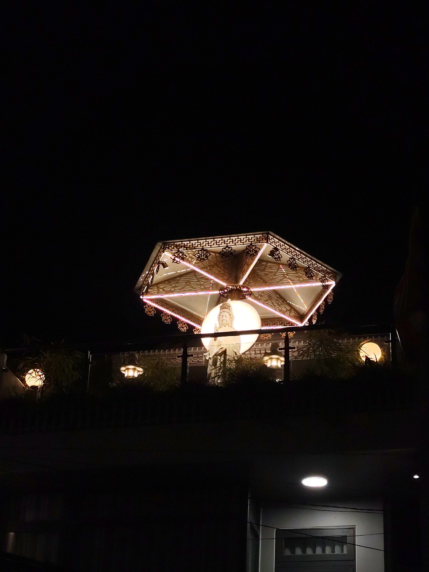 In the city of Phường Xuân Hương, Đà Lạt, Vietnam, a large, multi-faceted lantern hangs illuminated against the dark night sky. The lantern has a decorative, ornate metal frame with intricate patterns and is lit from within by a bright, circular light source that appears to be a statue or figure. Pink LED lights accent the edges of the lantern's structure. Below the main lantern, there are smaller, traditional-style lanterns and glowing orbs, some surrounded by foliage. The scene is set on what appears to be a balcony or elevated structure overlooking a darker area, possibly a street or lower level of a building. The overall impression is one of a festive or decorative night scene, likely in a public space or a well-lit establishment.