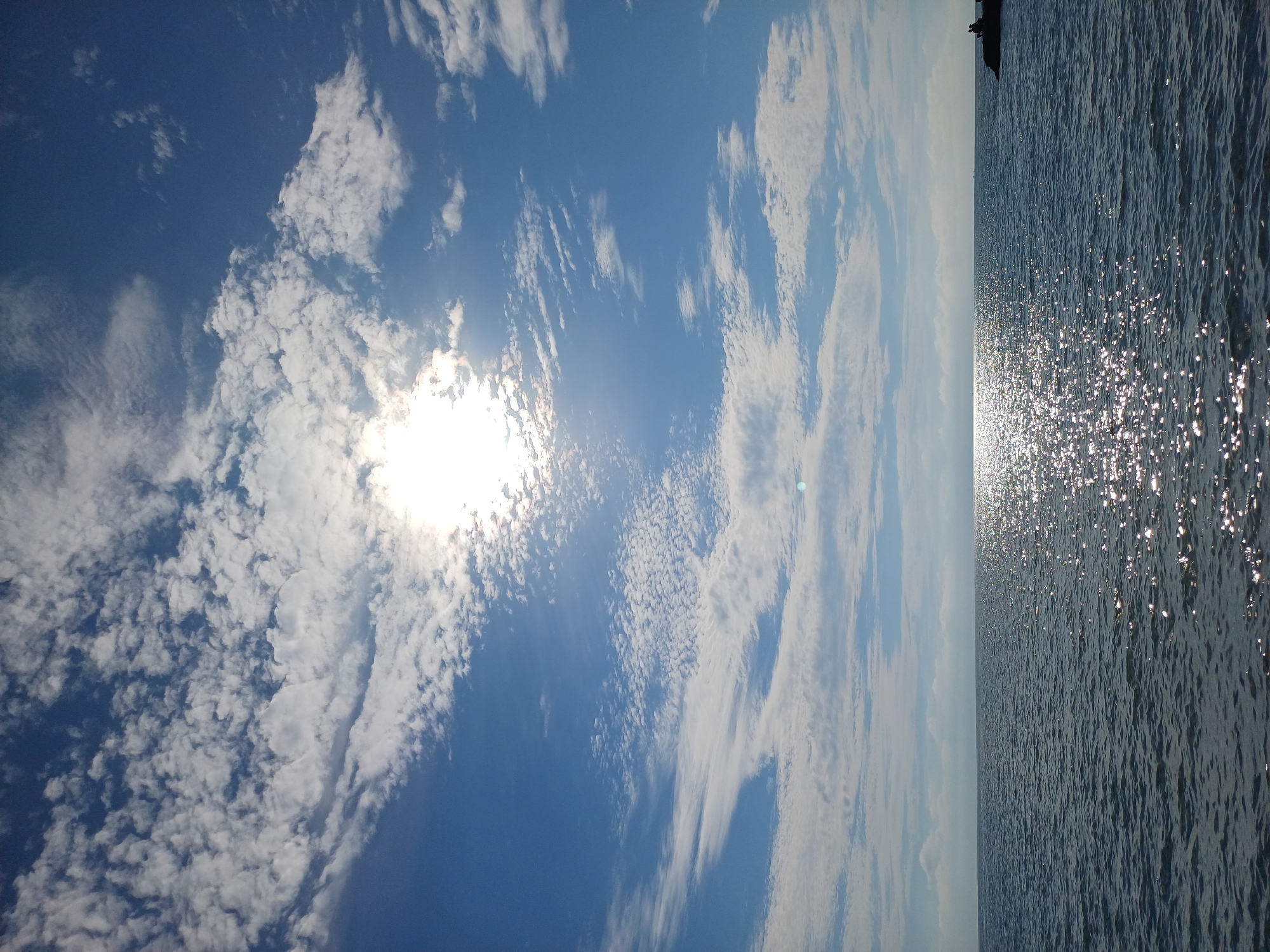 This is a vertical, full-frame shot of the sea and sky. The horizon line is low in the frame, with the sea occupying the bottom third and the sky the upper two-thirds. The sea is dark blue with small waves, reflecting the bright sunlight. The sky is a vivid blue, filled with a variety of white clouds, from thin, wispy cirrus clouds to larger, more textured cumulus clouds. The sun is partially obscured by clouds in the upper center of the frame, creating a bright glare. In the far right of the frame, on a rocky outcrop, two silhouetted figures can be seen sitting. The scene appears to be a coastal area, likely a beach or pier. Given the context, this image could be depicting the Black Sea coast in Sochi, Russia, during daylight hours.