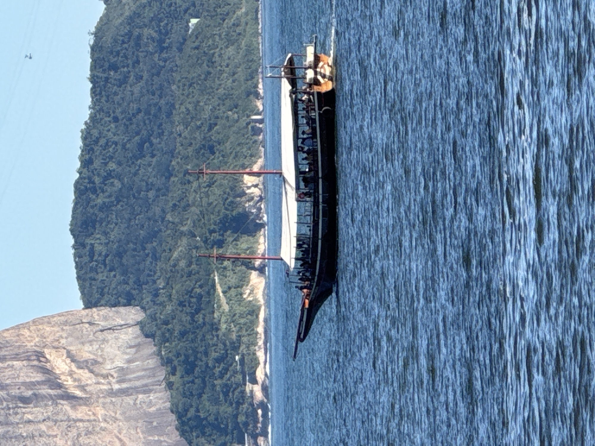 A tall ship sails on the blue water of the bay, with a mountainous, tree-covered landscape and a rocky cliff in the background. The ship, predominantly black with white sails partially furled, has a long deck with several people visible, some standing and some seated. One person is near the stern, wearing orange and white. The ship has two masts, one red and one darker, with rigging. The water is choppy, suggesting a breezy day. The time of day appears to be daytime, with clear skies. The location is Niterói, Brazil, with the distinctive landscape characteristic of the region. There is no visible text in the image.