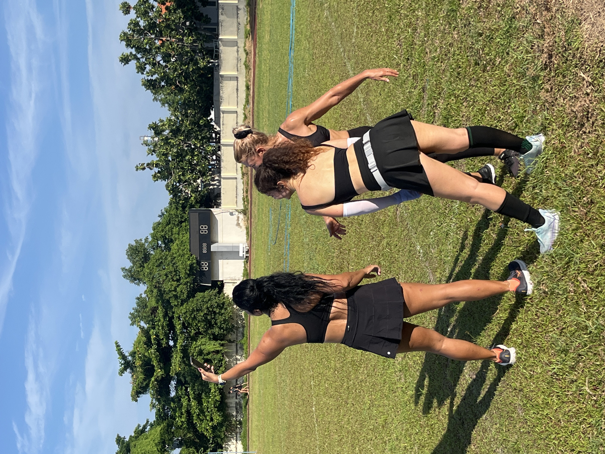 In a grassy outdoor stadium, three women are gathered. The woman in the foreground, facing away from the camera, is taking a selfie with her phone. She has long, dark hair and is wearing a black sports bra and a black pleated skirt. To her right are two other women, also wearing athletic attire. One, with blonde hair pulled back, wears a black sports bra and a black skirt. The other, in the mid-ground, has curly brown hair and is dressed in a black sports bra and a black skirt with a white belt detail. They are on a green field with a track visible in the background. A scoreboard displaying "88 88:88 88" is situated behind the women, and beyond that, trees and a white wall line the perimeter of the stadium. The sky above is blue with scattered white clouds. The scene appears to be set in the city of Phan Thiet Ward, Vietnam.