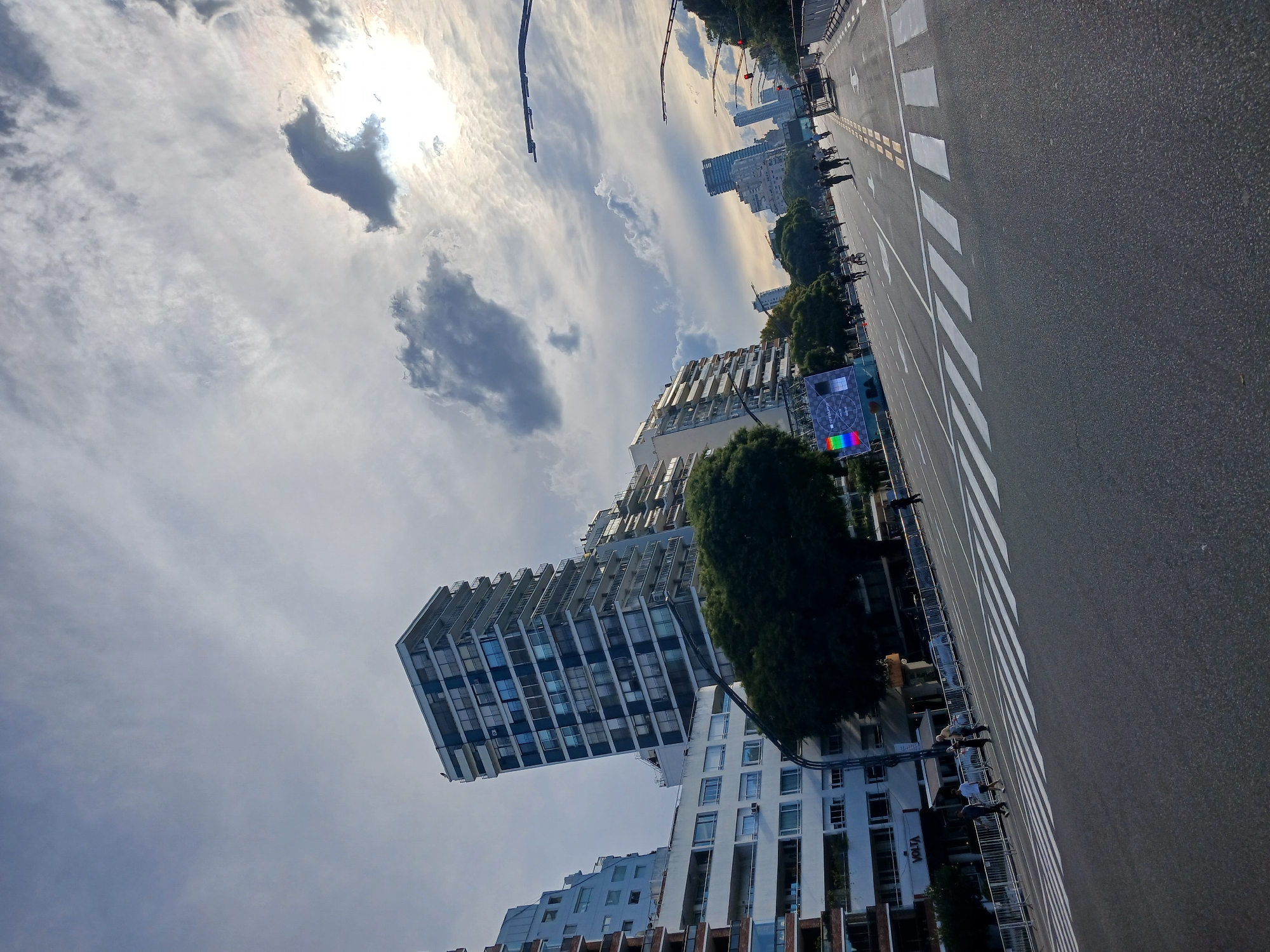 The image depicts a street scene in Buenos Aires, Argentina. The shot is taken at street level, looking towards the city skyline. Several tall buildings are visible, including a modern residential tower with multiple floors and a distinctive cantilevered design. In the foreground, a wide avenue stretches into the distance, marked by white crosswalk stripes and a grey asphalt surface. The sky is partly cloudy with the sun shining through. People are seen walking along the sidewalk adjacent to the street.