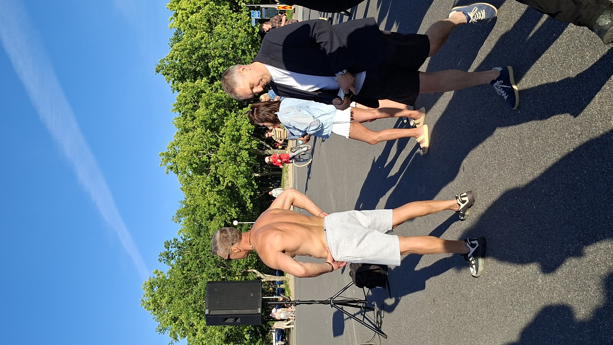 The image depicts an outdoor gathering on a sunny day with a clear blue sky and a few wispy clouds. Lush green trees line the background, suggesting a park or a tree-lined street. The pavement is visible in the foreground, marked with shadows cast by the sun.
The primary focus is on several individuals. In the foreground, a shirtless man with tanned skin is standing with his hands on his hips, wearing light grey shorts and sunglasses. To his left, a black speaker rests on a stand. Behind him, a man in a dark suit jacket, white t-shirt, and dark shorts is interacting with a woman. The woman is wearing a light blue denim jacket over a white top and white shorts. Further in the background, a person in a red jacket is visible near a bicycle, and other individuals can be seen amidst the trees.
The scene appears to be a public event, possibly a festival or an informal gathering, given the presence of a speaker and the mix of casual and more formal attire. The bright sunlight and long shadows suggest it is late afternoon. The overall mood seems relaxed and social. There is no visible text in the image. The location context provided is Tallinn, Estonia.
