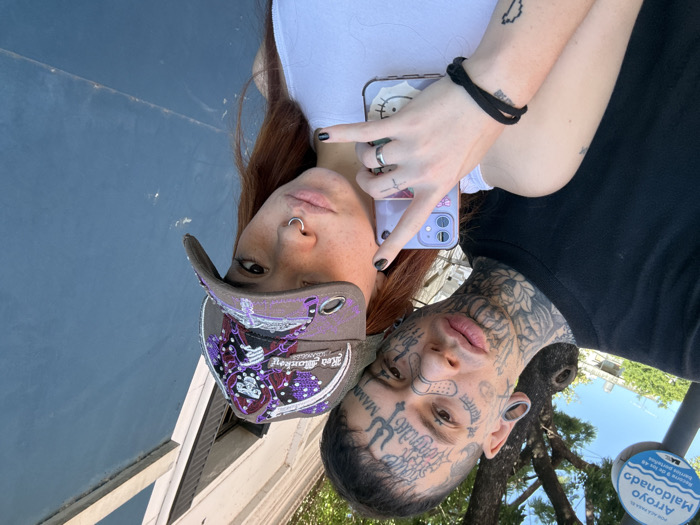 A selfie captures a pair of individuals. One person wears a ball cap and the other has extensive face tattoos. The person with the hat has red hair and a nose ring. The tattooed person has ear gauges, and a band-aid on their face. Both are wearing shirts; the person with the hat has a white shirt, while the tattooed person has a black shirt. One arm is visible, holding a phone with a Hello Kitty design. The sky and a sign are visible in the background.