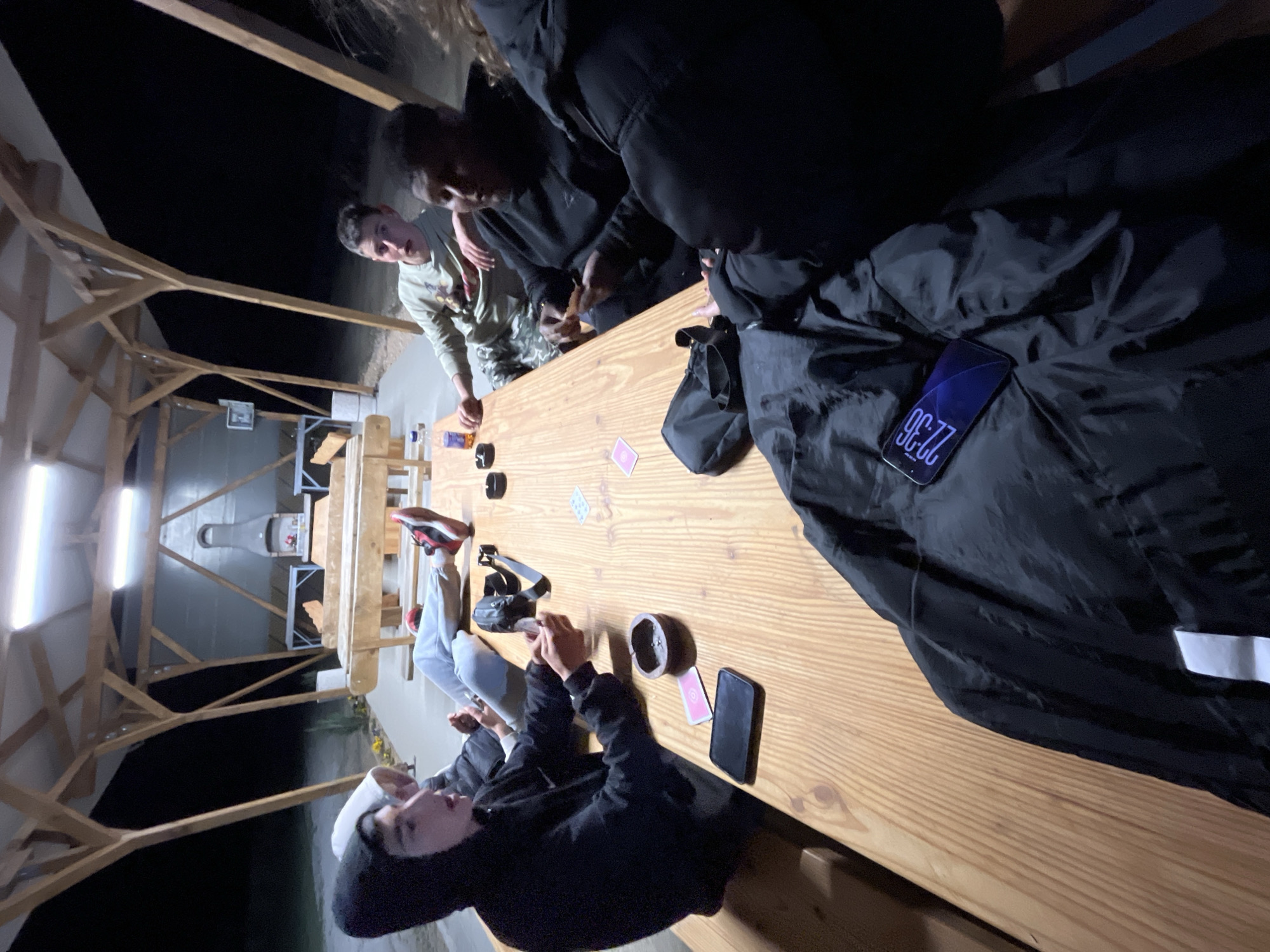 The image captures a casual nighttime gathering of young men around a long wooden table within an open-air wooden structure, likely a gazebo or shelter, in Karavelovo, Bulgaria. Bright fluorescent lights illuminate the scene from the wooden ceiling beams.
At least four individuals are visible. In the immediate foreground, a young man wearing a black hoodie and a white cap looks towards the camera. Further down the table, another young man in a green long-sleeve shirt (possibly military-style) with short hair is looking intently at something in his hands. Another person is largely obscured by a black puffer jacket draped over the table, on which a black smartphone displays the time "22:36". A fourth individual is seen from the waist down, with feet in light-colored pants and red-and-black sneakers resting on the table.
The wooden table holds several items, including two playing cards (a pink diamond and a green club are distinct), dark round objects (possibly ashtrays or small containers), a purple bottle, and two black smartphones. One of the phones clearly shows the time as 10:36 PM. In the background, beyond the table, a grill or smoker is visible. The surroundings outside the structure are dark, confirming the nighttime setting. The overall mood appears relaxed and social, suggesting friends enjoying a late-night hangout.