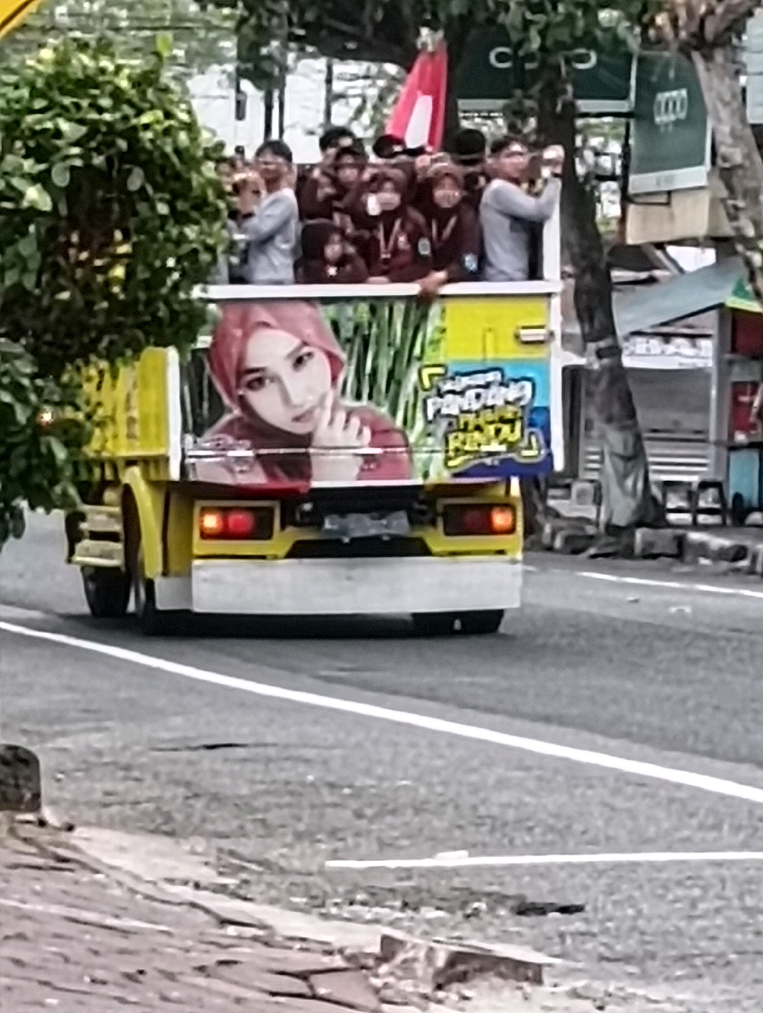 A low-resolution, grainy image shows a yellow truck driving on a paved road. The back of the truck features a large graphic of a woman wearing a pink hijab. Behind her, the text "PENDING CINTA RINDU" is visible in blue and yellow letters. Several people, some wearing maroon shirts, are visible standing in the back of the truck behind the graphic. The scene appears to be outdoors in an urban or suburban environment, with trees and buildings visible in the background. The presence of a red and white flag suggests a possible event or parade. The truck's taillights are illuminated red. The overall impression is a moving vehicle participating in a procession or gathering within a populated area.