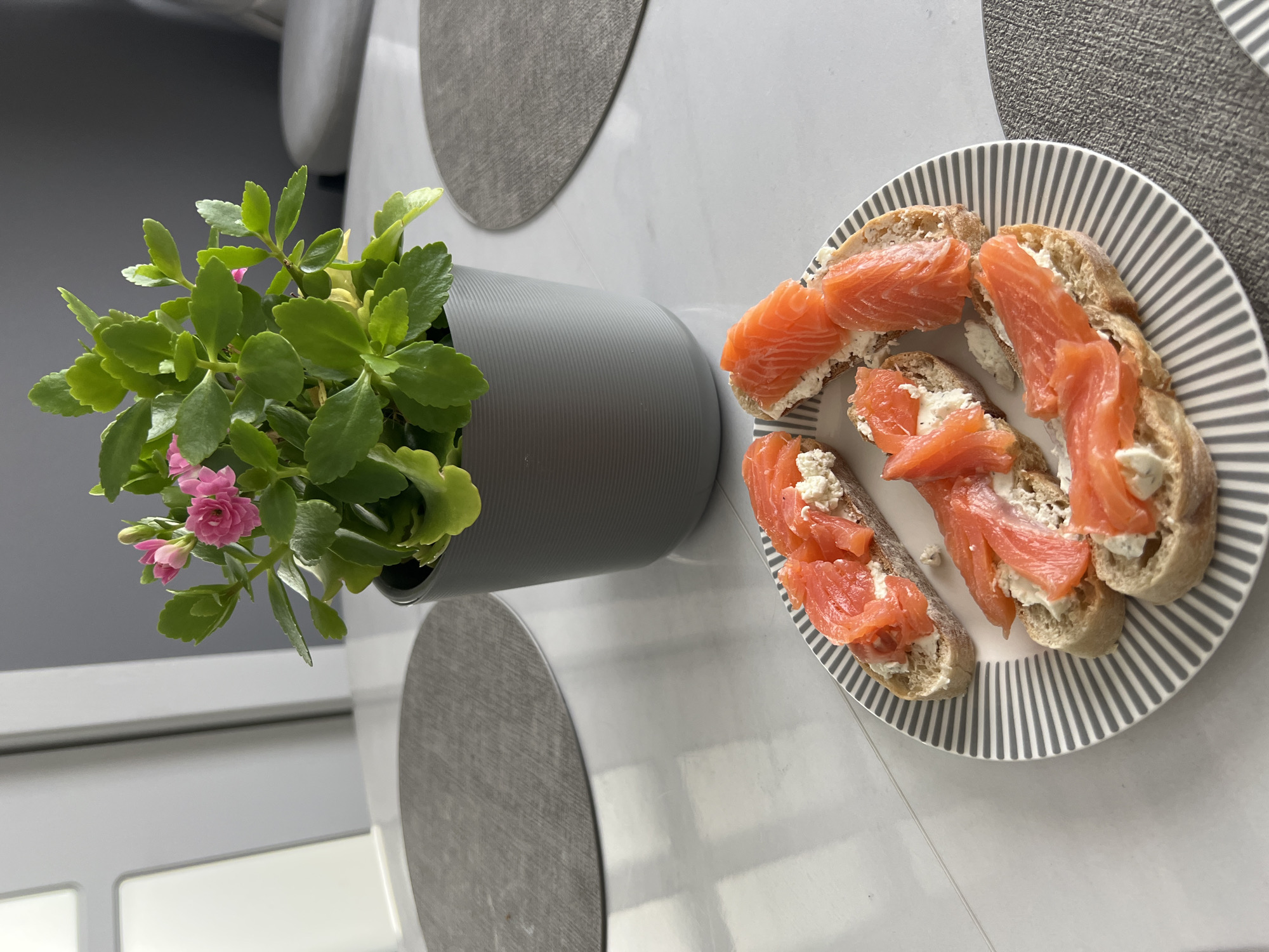 A top-down, medium shot captures a plate of open-faced sandwiches topped with cream cheese and slices of salmon. The plate is positioned on a light-colored, reflective surface, possibly a table with a marble-like pattern. To the right of the plate, a dark gray textured placemat is visible. In the background, a potted plant with green leaves and small pink flowers sits in a dark gray ribbed pot. The environment suggests an indoor setting, likely a dining area or kitchen, with hints of a doorway and a gray wall. There are no people visible in the image, and no specific identifiable landmarks that would place it in the city of Кулешовка, Russia.