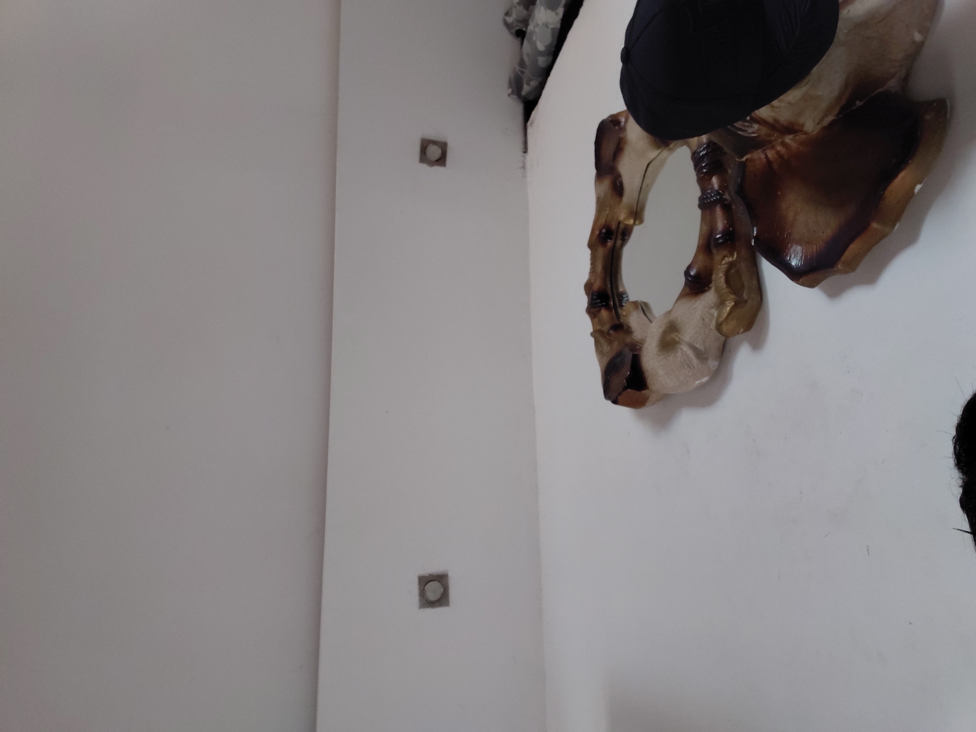 This image captures an indoor scene within a room in Santa Ana, Venezuela, showing a corner where two white walls meet. A decorative mirror with an ornate frame is mounted on the right wall. The frame exhibits carved, organic shapes, rendered in shades of dark brown and lighter gold or beige, giving it a rich, possibly wooden or faux wood appearance. A black cap or hat is perched on the upper edge of the mirror.
On the left wall, two small, square electrical outlets or cover plates are visible; each has a metallic, round button or center. Towards the top right corner of the image, where the walls converge, a small section of dark, possibly camouflage-patterned fabric is discernible. In the bottom right corner of the frame, a hint of dark hair suggests the reflection of the person taking the photograph. The setting is simple, characterized by plain white walls, and offers no specific indicators of the time of day, weather, or any text.
