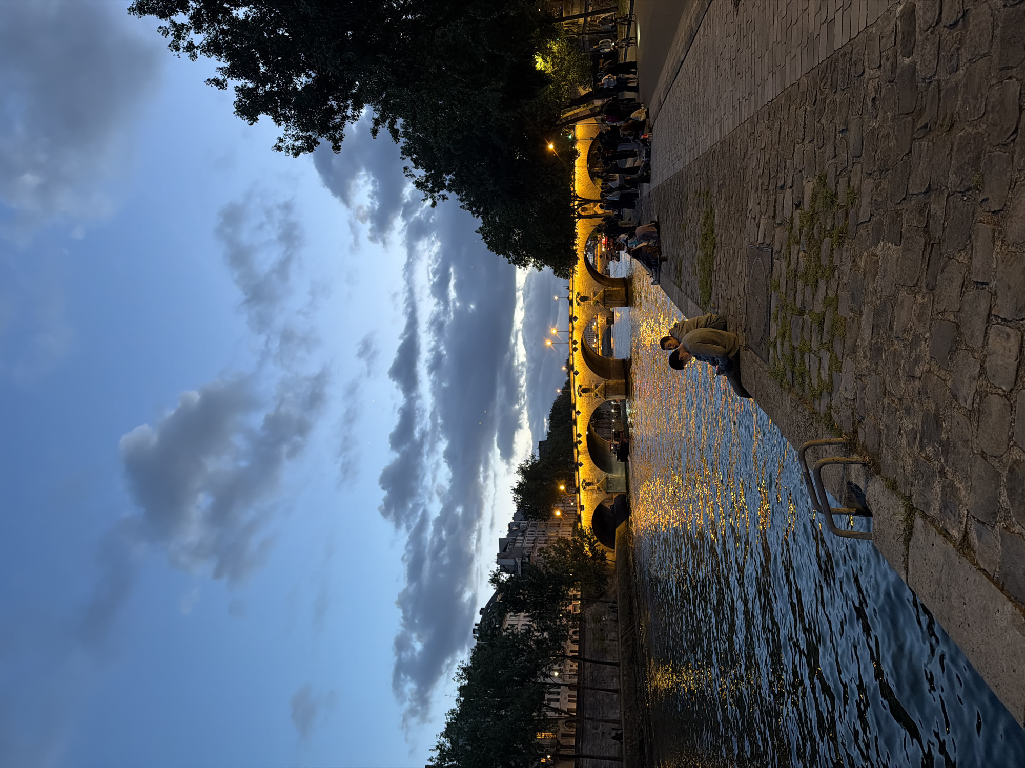This image captures an evening scene along the **River Seine in Paris, France**.
The sky above is a deep twilight blue, transitioning to a lighter hue towards the horizon, and is partially covered by scattered, dark grey clouds of varying density. Two faint, distant bright points are visible in the upper right sky.
The River Seine occupies the lower left portion of the frame. Its surface is dark blue with visible ripples, reflecting numerous elongated, shimmering golden-yellow lights from the bridge and adjacent streetlights. A small, dark boat is visible under the third arch from the left of the bridge.
A multi-arched stone bridge, prominently illuminated with golden-yellow lights, spans the river in the midground, its arches casting bright reflections onto the water.
On the far bank (left side of the river), a row of dark trees and classic multi-story Parisian buildings with numerous windows line the riverbank. Streetlights cast pools of light onto this distant embankment.
In the foreground, on the near bank (right side of the river), a wide stone quay or embankment is visible. It is constructed from large, irregular cobblestones with occasional patches of green vegetation. Two bent metal handrails extend into the water from the quay in the lower left corner.
Numerous individuals are present on this near bank. In the immediate lower left foreground, two men are seated on the edge of the quay, facing the river; one is looking downwards. Further along the quay, several small groups of people are seated directly on the cobblestones. To their right, a larger gathering of individuals stands and congregates on a wider, paved area alongside a row of trees that line the street. The trees are dark and largely silhouetted, with some branches catching light from overhead streetlamps. One person on a bicycle with an illuminated front light is visible on the far right of the street level. The overall scene suggests a relaxed social gathering along the river at dusk.