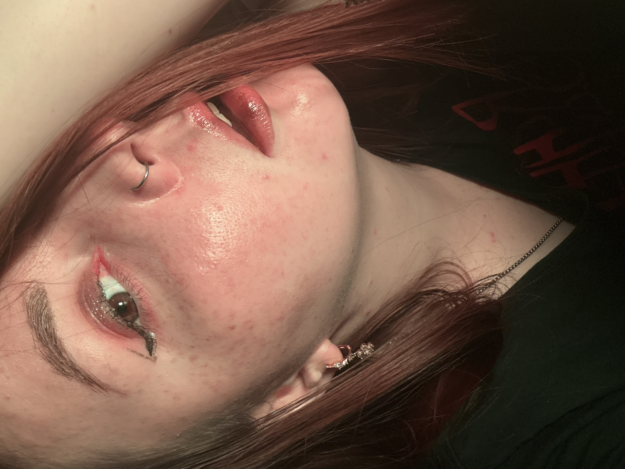 A close-up, tilted portrait depicts a fair-skinned individual with reddish-brown hair. The person has a nose ring on their left nostril and an earring on their left ear. The individual's left eye is adorned with glittery eyeliner. The lips are glossy and slightly parted. The person is wearing a black garment with red lettering on it. A silver chain is visible around the neck. The skin appears to have freckles and some redness. The setting and location of the image are not discernible, so it is not possible to say if this picture was taken in Izhevsk, Russia.