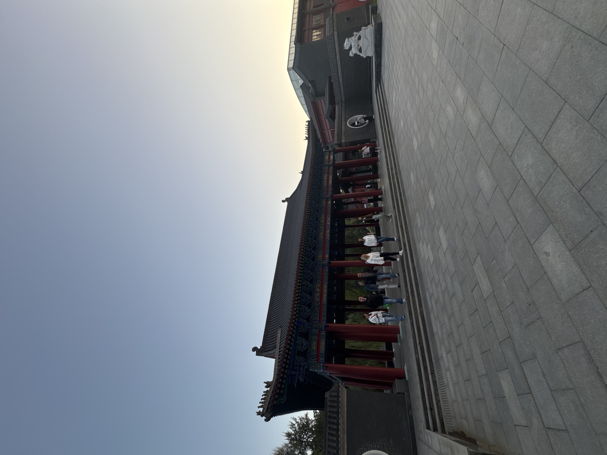 A wide-angle, low-angle outdoor shot captures a group of people walking on a paved courtyard in front of an ornate, traditional East Asian-style building with a dark, multi-tiered roof and red pillars. The sky is clear and a pale blue, transitioning to yellow near the horizon, suggesting either sunrise or sunset. The architecture features intricate decorative patterns under the eaves. To the right of the main building, a more modern structure with large windows is visible. A white stone sculpture of a mythical beast sits on a low wall in the foreground. Several individuals are walking from left to right across the image, some in casual clothing, others in more formal attire. The ground is paved with large, rectangular grey stones.