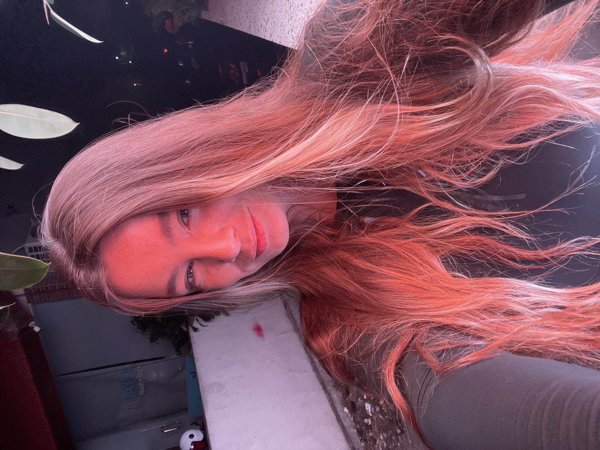 A young woman with long, wavy, blonde hair dyed with reddish-pink highlights is shown in a selfie. She is wearing a dark, long-sleeved shirt. The lighting in the image is predominantly red, casting a warm glow on her face and hair. Her left eye is visible, looking towards the camera. In the background, to the left, is a white wall with some graffiti and what appears to be a sign that reads "7 DAYS WASH DRY". Behind her, the background is dark, with some blurred greenery and lights, suggesting an outdoor nighttime setting. To the right of the image, a black vehicle is partially visible. The overall scene suggests the woman is outdoors at night, possibly in a street or an area with some commercial establishments like a laundromat, given the sign. The specific location in North Kuta, Indonesia, cannot be determined from the visual information alone.