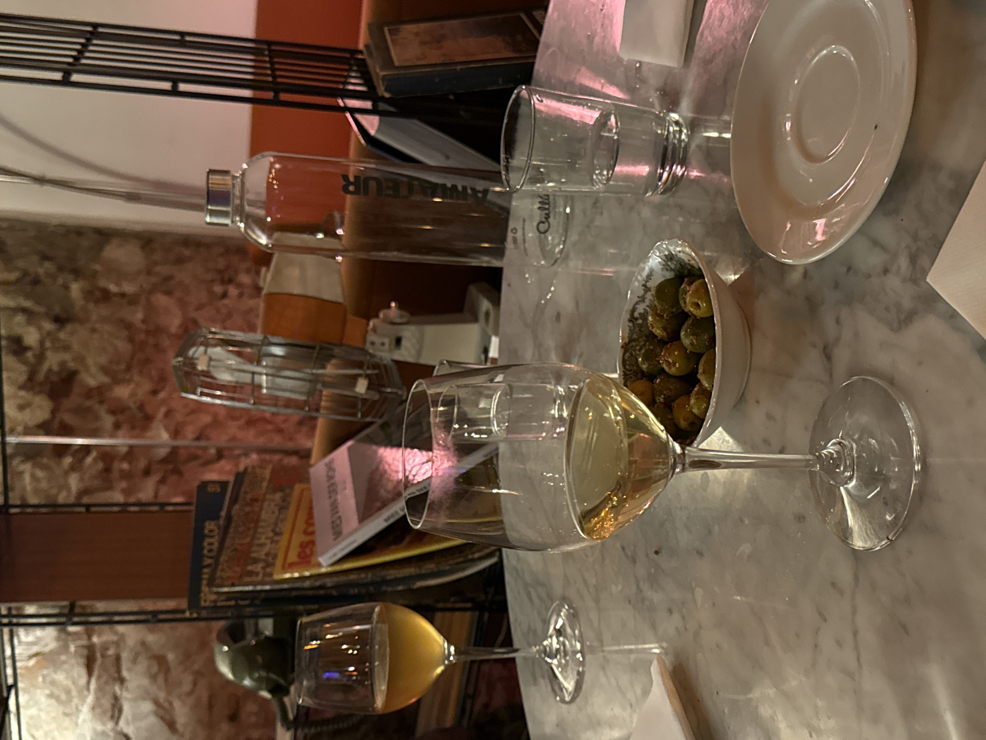 The image shows a still life on a marble table, likely in a bar or restaurant. There are two wine glasses, one filled with white wine and the other with a beverage of a golden hue. A small bowl of olives is positioned between the glasses. A glass water bottle labeled "Amateur" and a drinking glass with "Culle" visible are placed near a white plate. In the background, a shelf is visible holding books and other items. Based on the language of the books and the style of the setting, it is likely that this photograph was taken in the city of Barcelona, Spain. No people are visible in the frame.