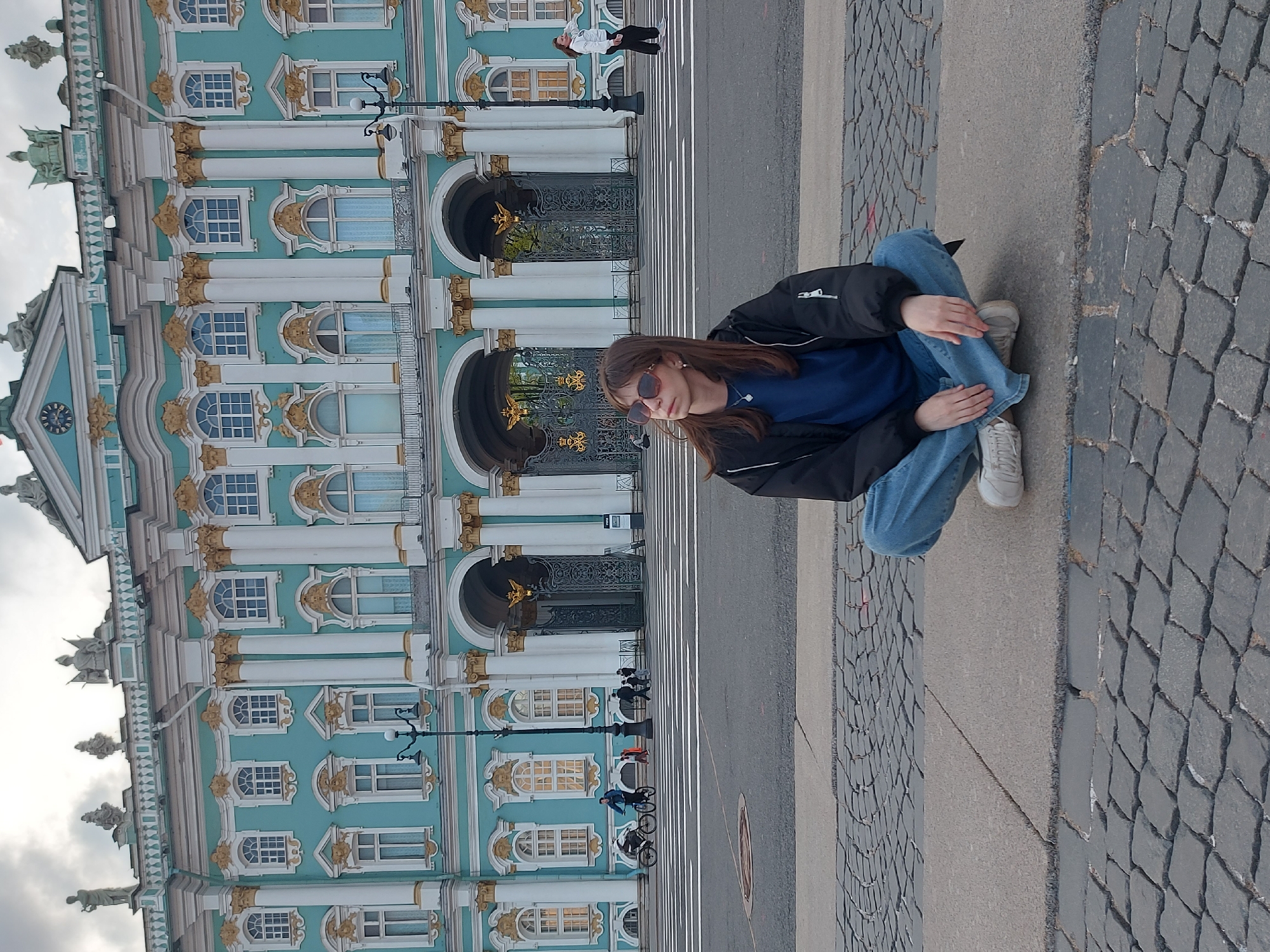 A vertical shot shows a person sitting on the pavement, with the Winter Palace in the background. The person is wearing blue jeans, a black jacket, a blue shirt, and white sneakers. They are sitting cross-legged and appear to be looking down. The Winter Palace, a blue and white baroque-style building with gold accents, stands tall in the background. The street in front of the palace is visible, with white lines marking pedestrian lanes. People on bicycles are seen on the road. The pavement where the person is sitting is made of grey stone blocks. The photo was taken in Saint Petersburg, Russia, with the Winter Palace serving as a notable landmark.