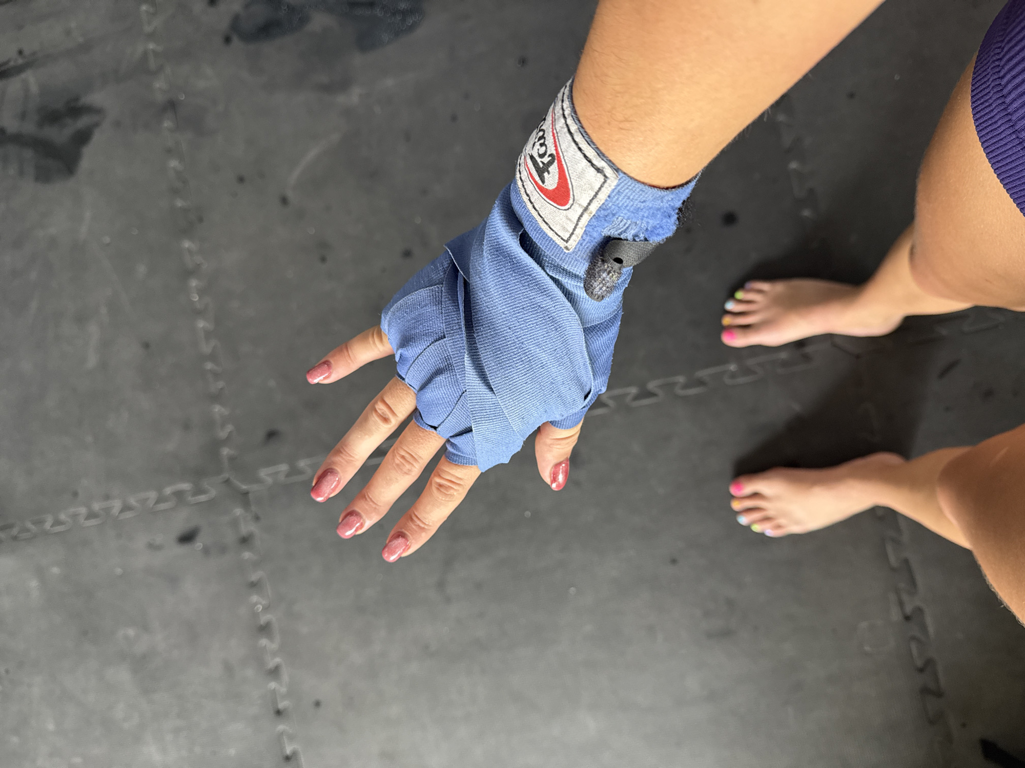An overhead shot shows a woman's hand wrapped in blue boxing wraps, with the brand "Fairtex" visible on the wrist. Her fingernails are painted a light pink color. The photo also captures part of her legs and feet, the toes painted with different colors. The scene unfolds on a textured gray surface, possibly interlocking mats commonly found in gyms. While the location in Tibubeneng, Indonesia is mentioned, there is no explicit information within the image to verify it.