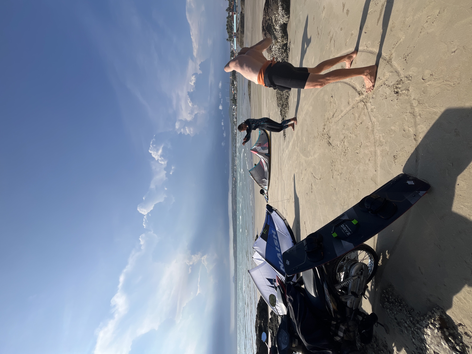 This is a wide-angle, outdoor photograph taken on a sandy beach in Mũi Né, Vietnam, under a bright blue sky with scattered white clouds and dramatic shafts of sunlight. In the foreground, a black kiteboard stands upright, leaning against a parked motorcycle which is covered with dark fabric. To the left of the motorcycle, rocks and shells line the edge of the sand.
In the mid-ground, two people are present on the beach. A man with a bare torso and dark shorts is facing away from the camera, with his arms raised and bent, appearing to be stretching or performing a dynamic movement. Further back, a woman in a dark long-sleeved rash guard and leggings is also facing away, her arms extended to the sides. Several kiteboarding kites are laid out on the sand between the two individuals and the motorcycle.
The background shows the turquoise ocean with moderate waves and a distant shoreline with buildings, palm trees, and some wind turbines, characteristic of the Mũi Né coastline. The overall scene depicts preparations or a break from kiteboarding activities.