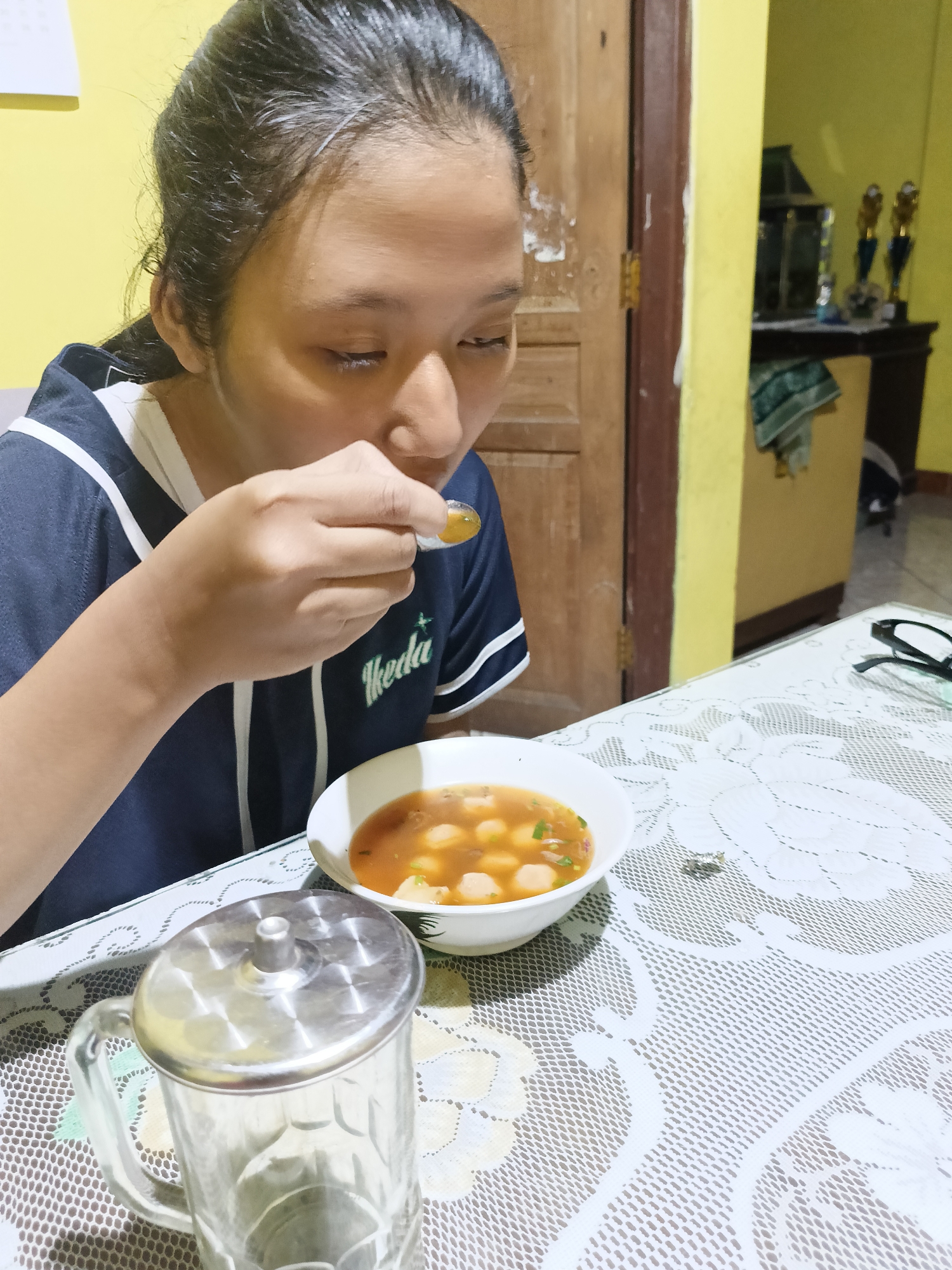 In Sukorini, Indonesia, a young woman with dark hair pulled back is seated at a table, eating a bowl of soup. She is wearing a navy blue shirt with white trim and white lettering that reads "Ikeda". A spoon is in her mouth, holding a spoonful of the soup, which appears to contain meatballs and green herbs. In the foreground, a clear glass mug with a metal lid sits on a white lace tablecloth. The background shows a yellow wall, a wooden door, and a shelf with trophies.