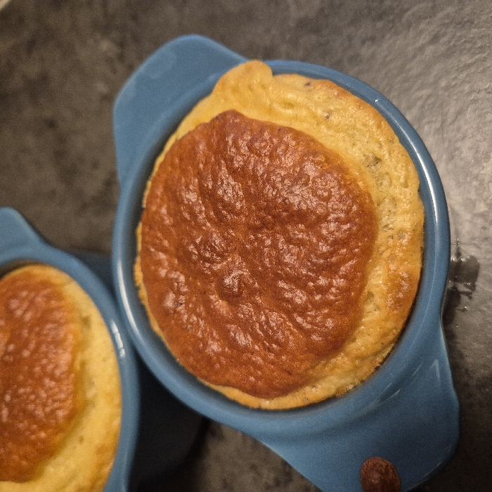 Two individual portions of a baked dish are presented in blue, oval-shaped ceramic ramekins with small handles. The dish features a prominently risen, deeply golden-brown, and crusted top surface over a lighter, golden-yellow, aerated base. The foreground ramekin is in sharp focus, with a second similar ramekin blurred in the background, positioned to the upper left. Both ramekins rest on a dark gray or black countertop with a subtle textured pattern. No individuals or specific actions are depicted. The scene is situated in Strasbourg, France.