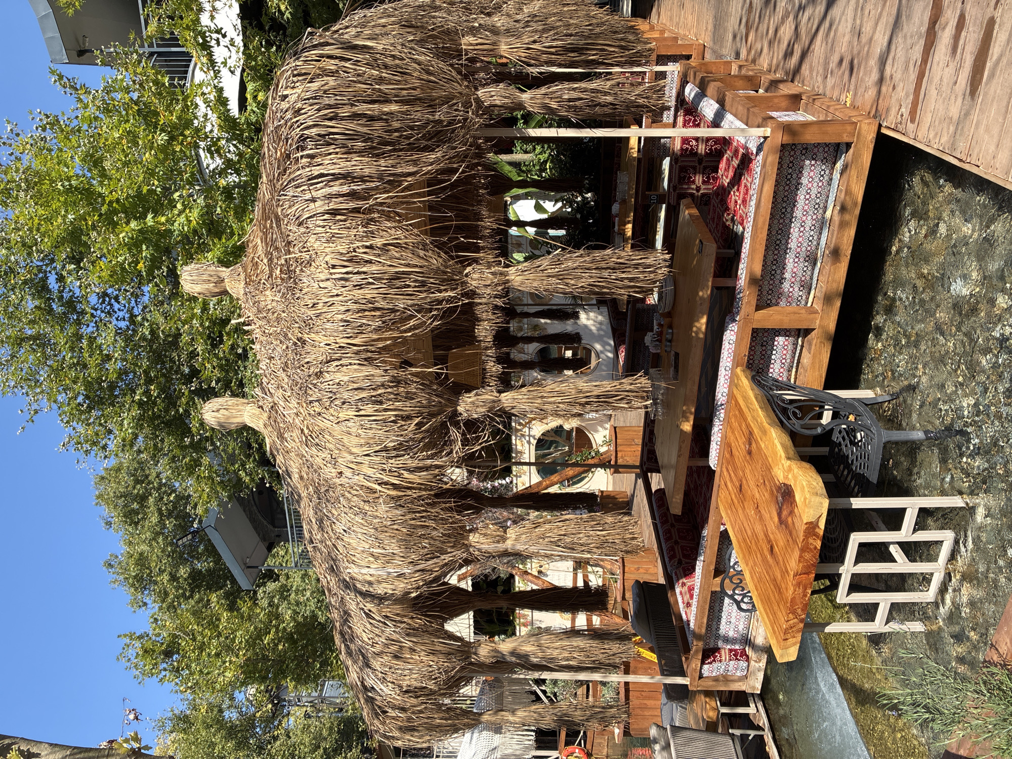 The image shows an outdoor seating area for a restaurant or cafe, situated next to a body of water, likely a canal or river, in Alanya, Turkey. The seating is arranged on a wooden platform that extends over the water. A prominent thatched roof provides shade for the seating area, which consists of wooden benches with colorful patterned cushions. Several wooden tables are placed around the benches, with one individual table made of a large, live-edge wood slab and black metal chairs positioned over the water. The environment is lush and green, with trees visible in the background, and the sky is clear and blue. The overall scene suggests a tranquil and scenic dining experience.