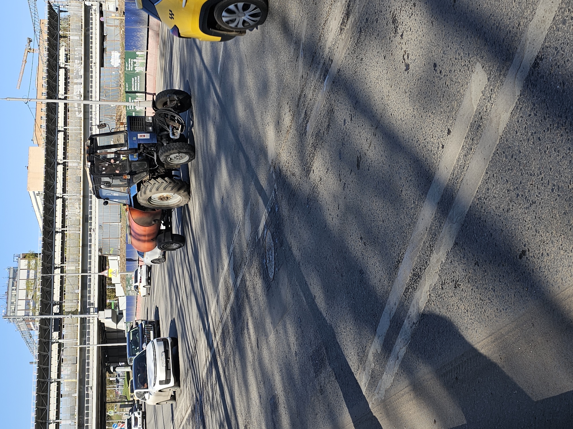 The image captures a street scene in Moscow, Russia, featuring various vehicles and a construction site.
Foreground: A grey asphalt road occupies a significant portion of the image. White lane markings are visible, indicating traffic direction. The road surface shows signs of wear.
Vehicles: A yellow taxi is partially visible at the top, with a visible wheel. Further down the street, a blue tractor is positioned. A row of cars, primarily white, is parked alongside the road.
Construction Site: A multi-story building under construction forms a backdrop on the left. Construction cranes and scaffolding are visible.
Environment: The sky is clear and blue. Shadows cast by nearby structures are prominent on the road surface.