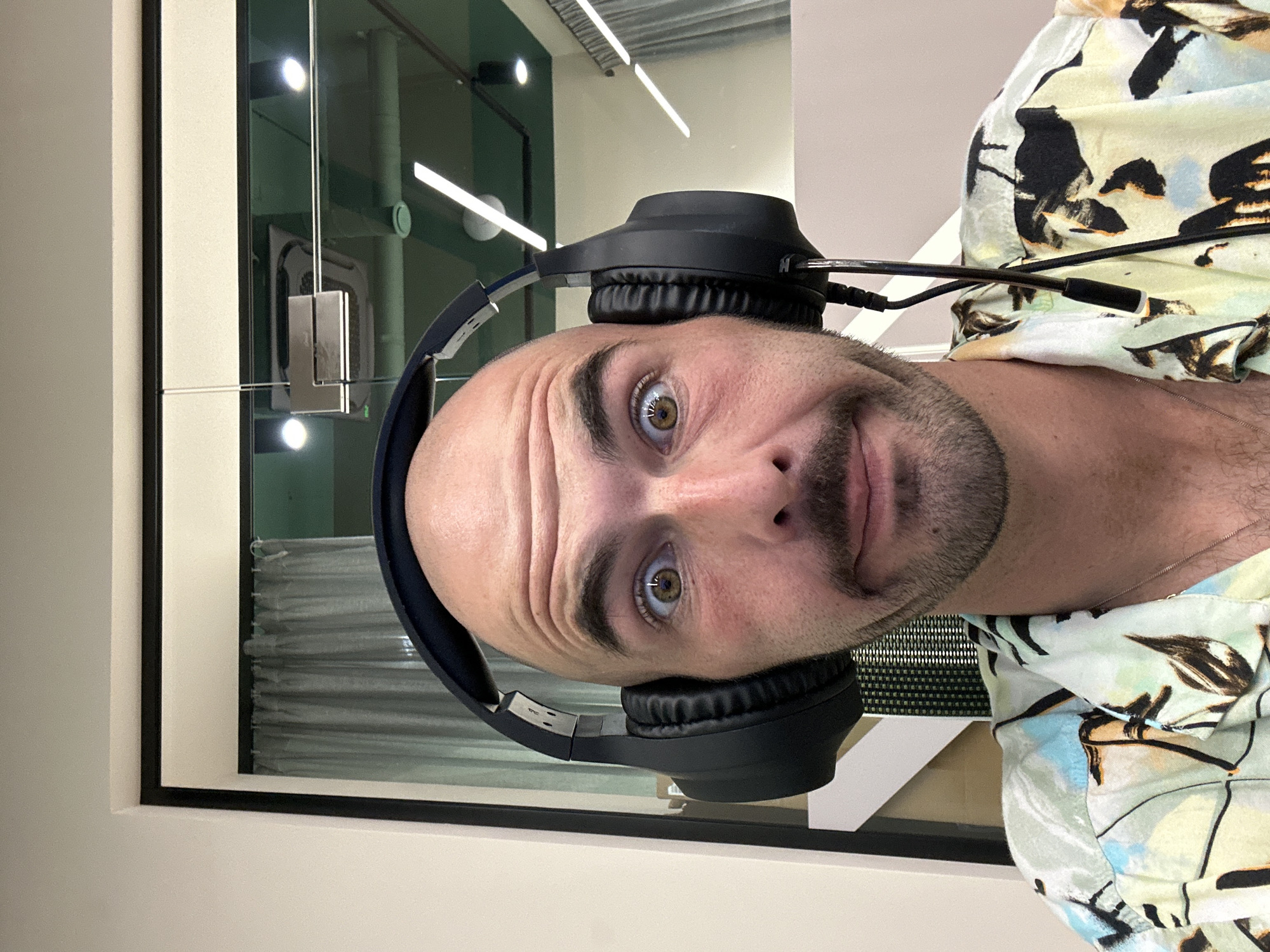 A man is wearing black headphones and a light-colored shirt with a floral pattern. He appears to be indoors, possibly in an office setting, as suggested by the window showing a building interior with ceiling lights. The location in Budva, Montenegro, is not discernible from the visual data.