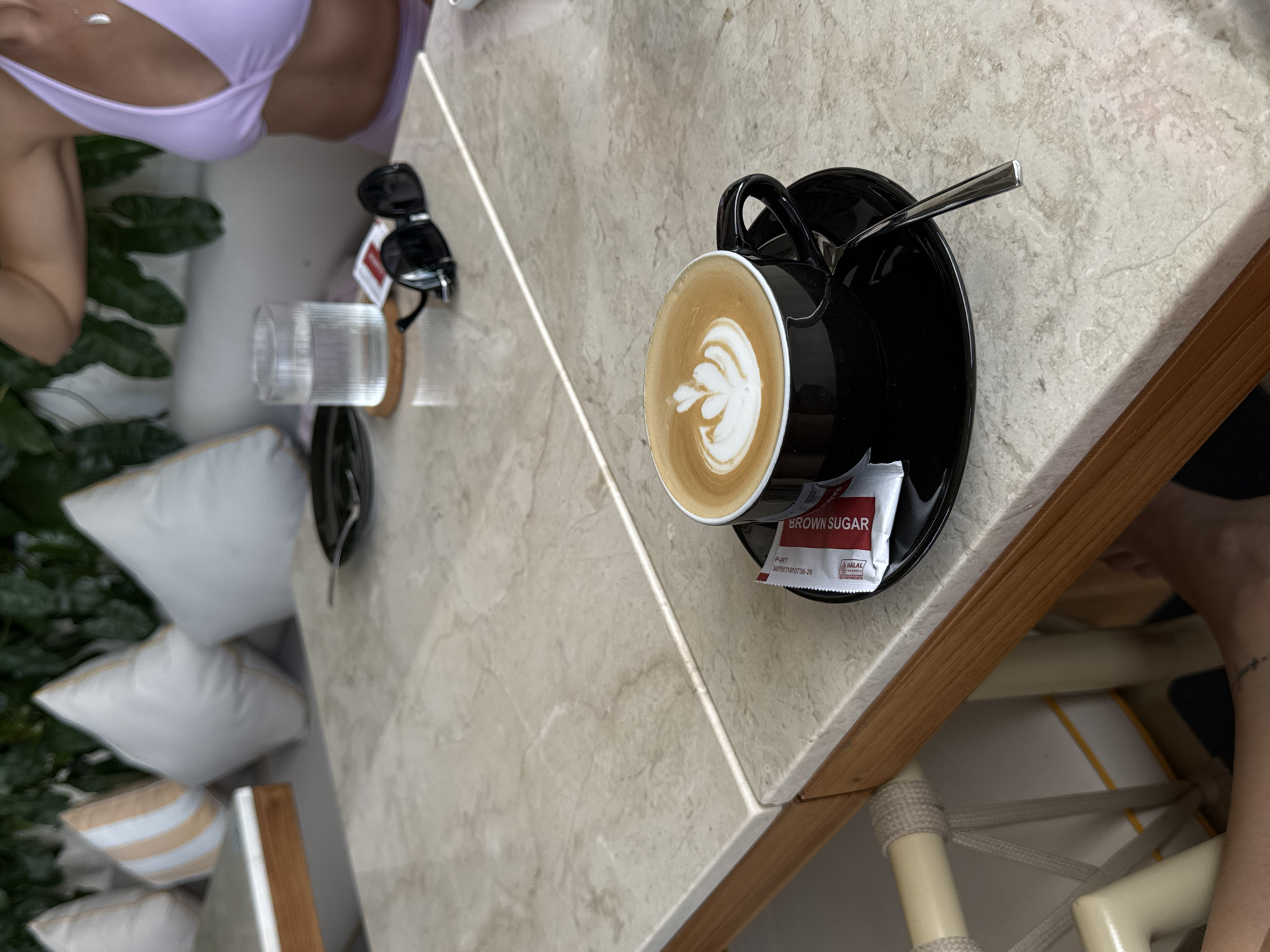 An overhead, close-up shot captures a black mug of latte art coffee resting on a black saucer on a light marble table. Next to the mug is a packet of brown sugar and a spoon. In the background, a glass of water and sunglasses sit on the table. The scene appears to be set outdoors, possibly at a cafe in North Kuta, Indonesia, indicated by the tropical foliage and relaxed ambiance. A person wearing a purple bikini is partially visible in the upper right corner, suggesting a beach or poolside setting.