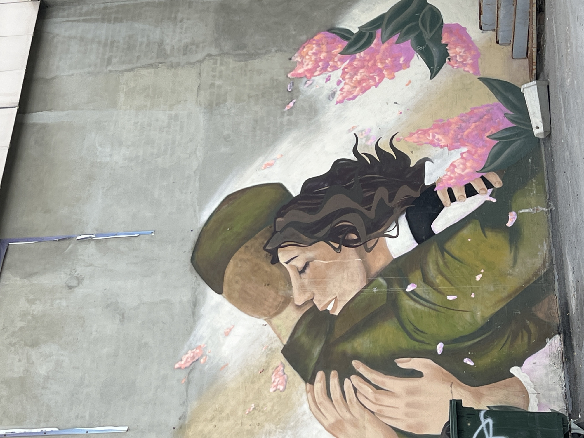 A mural on a concrete wall in Chelyabinsk, Russia, depicts a woman in a dark-haired updo embracing a man wearing a green military hat and coat. Pink flowers and green leaves are painted above them. The wall is mottled grey with signs of wear and repair. A small green object is visible in the bottom right corner. Patches of blue tape are present on the wall to the left of the mural.