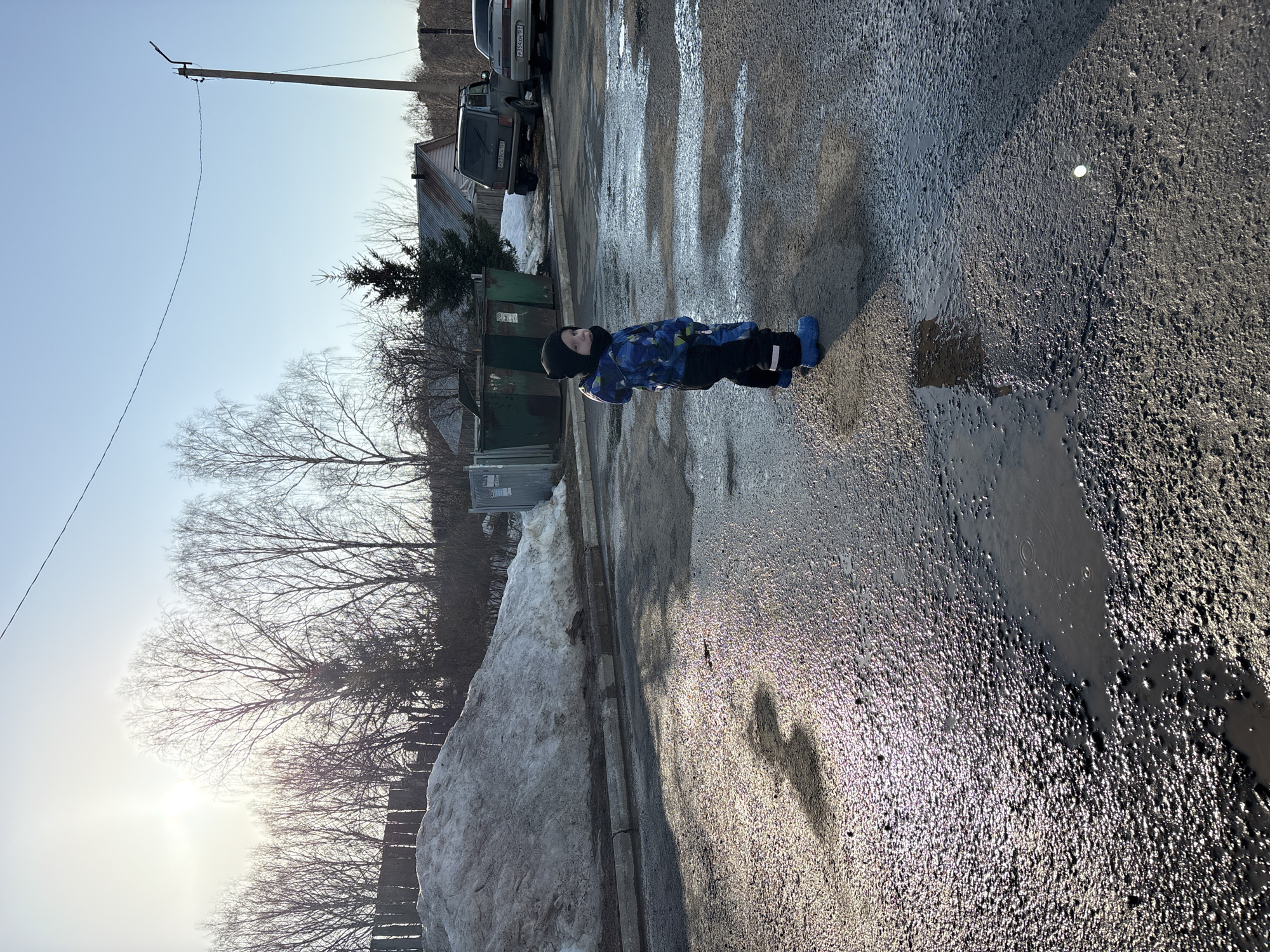 The image captures a single young child standing on a wet, uneven asphalt road in Chaikovsky, Russia, under bright daylight. The child wears a black beanie, a blue and black patterned winter suit, and blue rubber boots, positioned facing the right of the frame with their head turned to look left. The road surface is visibly worn, covered with numerous puddles and wet areas that reflect the sunlight. To the left, a large pile of melting snow is situated behind a concrete curb and a wooden fence. Bare deciduous trees are silhouetted against a bright, hazy sun in the upper left background. On the right, a wooden utility pole with overhead wires extends into a clear blue sky. Two vehicles are parked in the background on the right: a dark-colored SUV (license plate M039HM 159, indicating Perm Krai) and a lighter-colored sedan (license plate P354KH 159, also Perm Krai). Two large green industrial-style trash bins are located near the parked cars. A coniferous tree stands between the trash bins and a building with a corrugated metal roof in the distant background. The overall environment suggests late winter or early spring conditions, characterized by melting snow and clear weather.