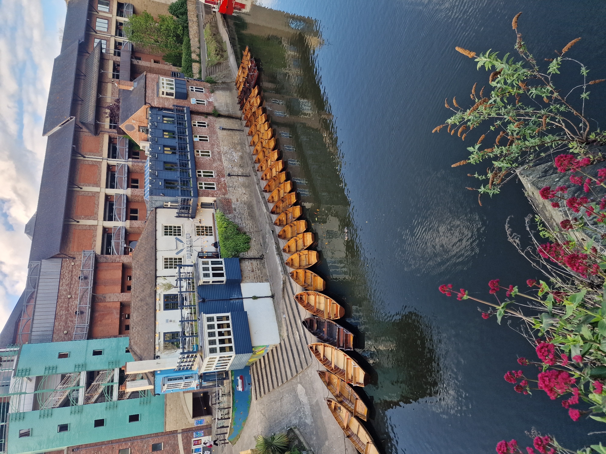 The image shows a high-angle, riverside view in Durham, United Kingdom. A line of rowing boats (likely punts) is docked along the concrete riverbank. The river is dark and reflects some of the surrounding buildings. On the left side of the image are buildings with a mixture of architectural styles, varying in color from brick to teal. Some of these buildings appear to be restaurants with outdoor seating. A staircase leads down to the river. In the foreground, some flowering plants with red and brown flowers hang over the edge of the riverbank. No people are prominently visible. The sky is visible with partial clouds.