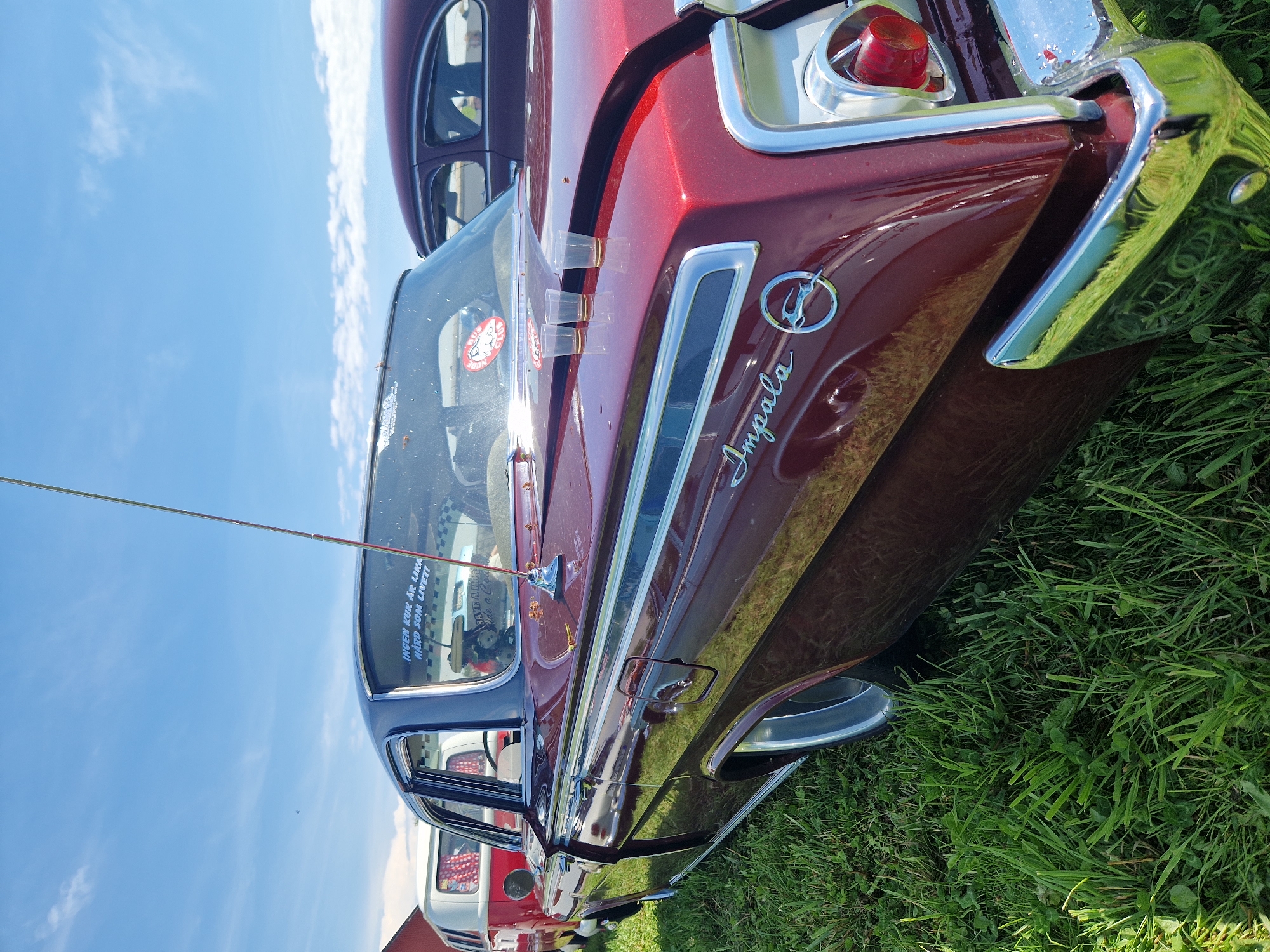 The image depicts a row of classic cars parked on a grassy field under a bright blue sky with scattered clouds. The primary focus is on the rear of a maroon Chevrolet Impala, viewed from a low angle. The car features chrome accents, a prominent "Impala" emblem, and a distinctive circular logo. Three clear plastic cups, appearing empty, are placed on the trunk lid of the Impala. To the left of the Impala, a white Volkswagen bus with red accents is partially visible. In the background, another dark-colored sedan is parked. The scene is outdoors, with lush green grass and a clear sky suggesting a daytime setting. The presence of multiple vintage vehicles indicates a car show or similar event. While the prompt mentions Edenberga, Sweden, there are no specific visual cues within the image to confirm this location.