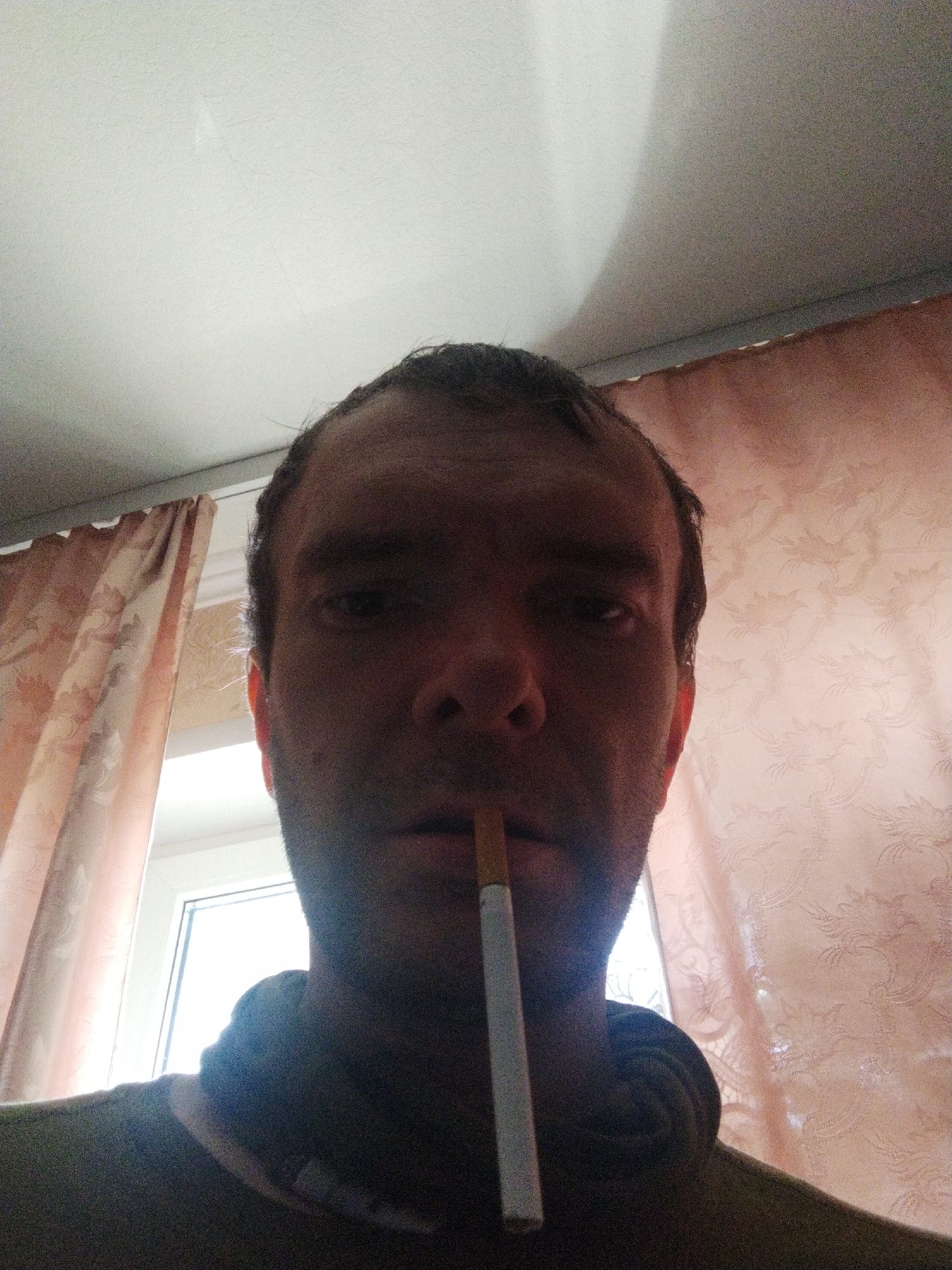 A man is holding an unlit cigarette in his mouth, photographed from a low angle. He has short, dark hair and appears to be indoors, with a window and patterned curtains visible in the background. The ceiling is also visible. The man's face is partially shadowed. The location provided is Kramatorsk, Ukraine, but there is nothing visible in the image to confirm this.