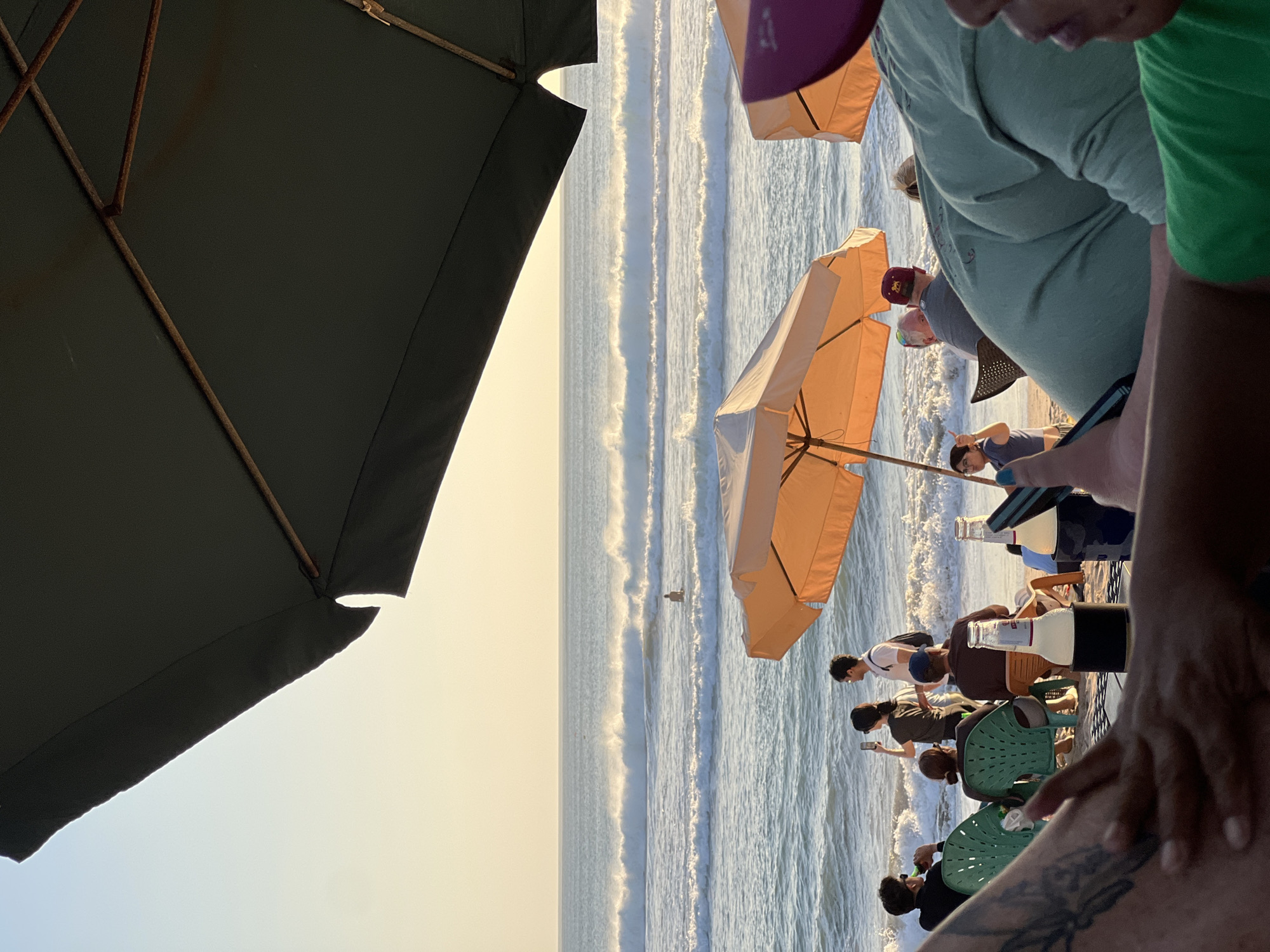 The image depicts a beach scene in Kuta, Indonesia, likely during late afternoon or early evening, judging by the soft, warm light. Two large umbrellas, one dark gray and the other orange, are prominently featured, casting shadows and providing shade. The dark gray umbrella is in the foreground, angled to cover a portion of the view. The orange umbrella is smaller and further back, situated closer to the water.
Several people are visible in the image, enjoying the beach. Some are seated on chairs, including a group of individuals under the orange umbrella. One person is standing by the water, appearing to be a surfer or swimmer. Another group is standing on the sand, with one person taking a photo with their phone. Two bottles of drinks are visible on a surface in the foreground.
The ocean is characterized by significant waves breaking near the shore, creating white foam and ripples. The water appears a deep blue-green. The sky is a pale yellow-orange, indicating either sunrise or sunset. The sand is light brown. The overall atmosphere suggests a relaxed and leisurely environment.