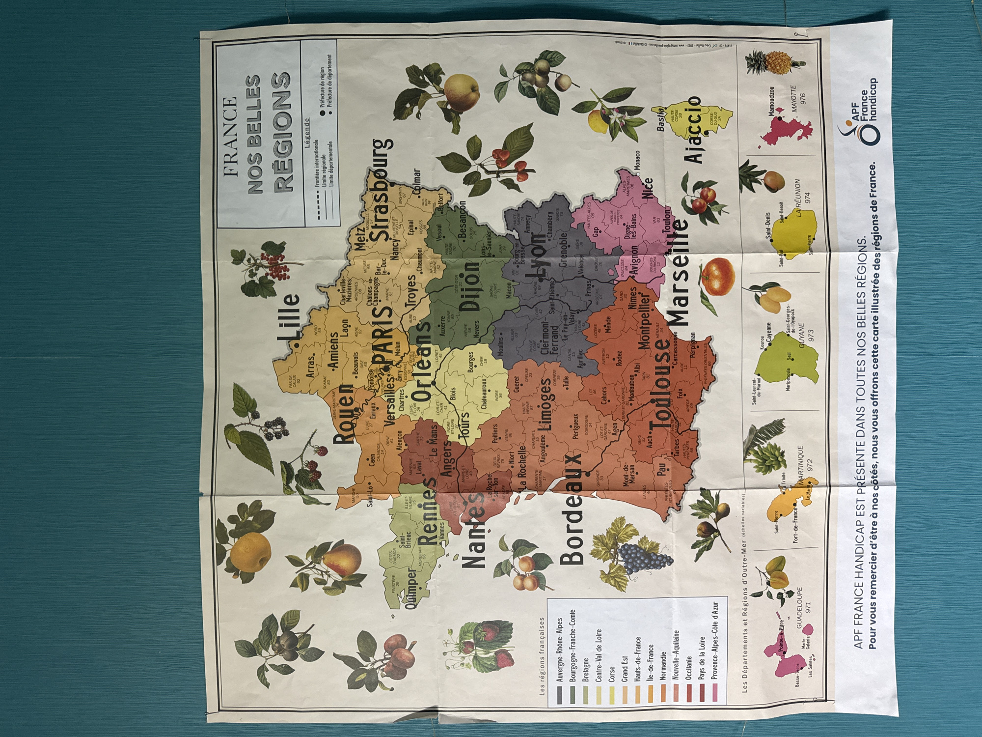 The image is a map of France and its overseas territories, titled "FRANCE NOS BELLES REGIONS" (France Our Beautiful Regions). The map highlights the different regions of France using various colors, a color-coded key is provided at the bottom left. International, regional, and departmental boundaries are indicated with dotted lines. Cities, identified with a dot, are labeled, with the regional capitals bolded. Fruits and berries are distributed around the map, each associated with a specific region. Below the map are located depictions of the overseas departments and regions of France, complete with illustrations of local produce. The bottom of the map features text: "APF FRANCE HANDICAP EST PRÉSENTE DANS TOUTES NOS BELLES RÉGIONS. Pour vous remercier d'être à nos côtés, nous vous offrons cette carte illustrée des régions de France." (APF FRANCE HANDICAP IS PRESENT IN ALL OUR BEAUTIFUL REGIONS. To thank you for being by our side, we offer you this illustrated map of the regions of France.). The upper right corner includes the APF France Handicap logo.
