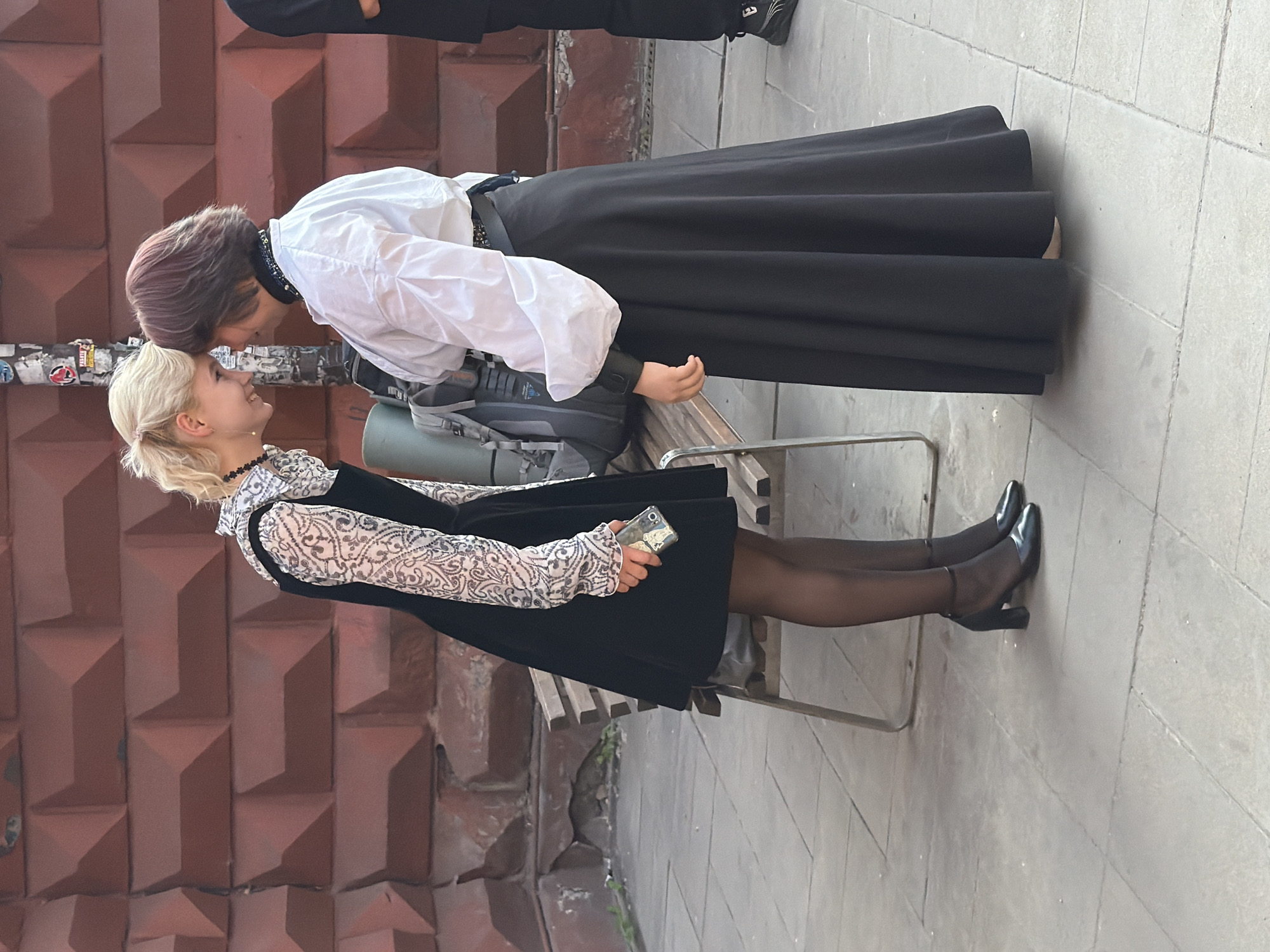Two young women are embracing, with their foreheads touching. One woman has short blonde hair, is wearing a black dress with a white, patterned top, black tights, and black heeled shoes. She is holding a smartphone in her right hand. The other woman has dark hair with purple streaks, is wearing a white long-sleeved shirt, a black skirt, and a black collar. She is leaning forward to touch foreheads with the other woman. They are standing outdoors on a paved sidewalk next to a red brick wall with geometric patterns. A backpack is visible behind the women. The scene appears to be set during the daytime.