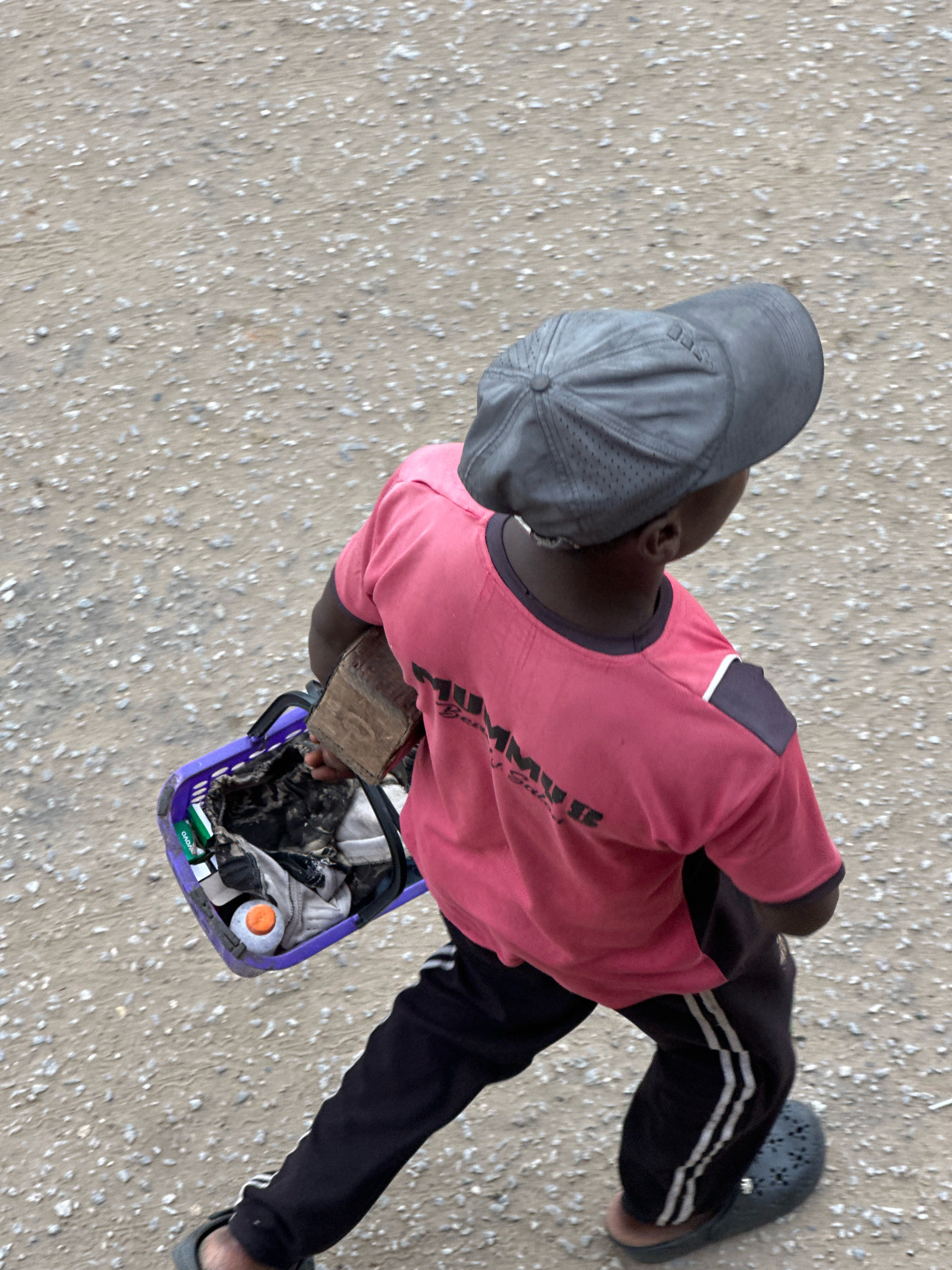 A person wearing a pink t-shirt and black pants with white stripes walks on a dirt path in Ajara, Nigeria. The person is carrying a purple basket in their left hand. The basket contains various items, including a water bottle with an orange cap and some clothing. The person is also holding a piece of wood. They are wearing a grey baseball cap and dark-colored sandals. The ground is covered in small stones and dirt.