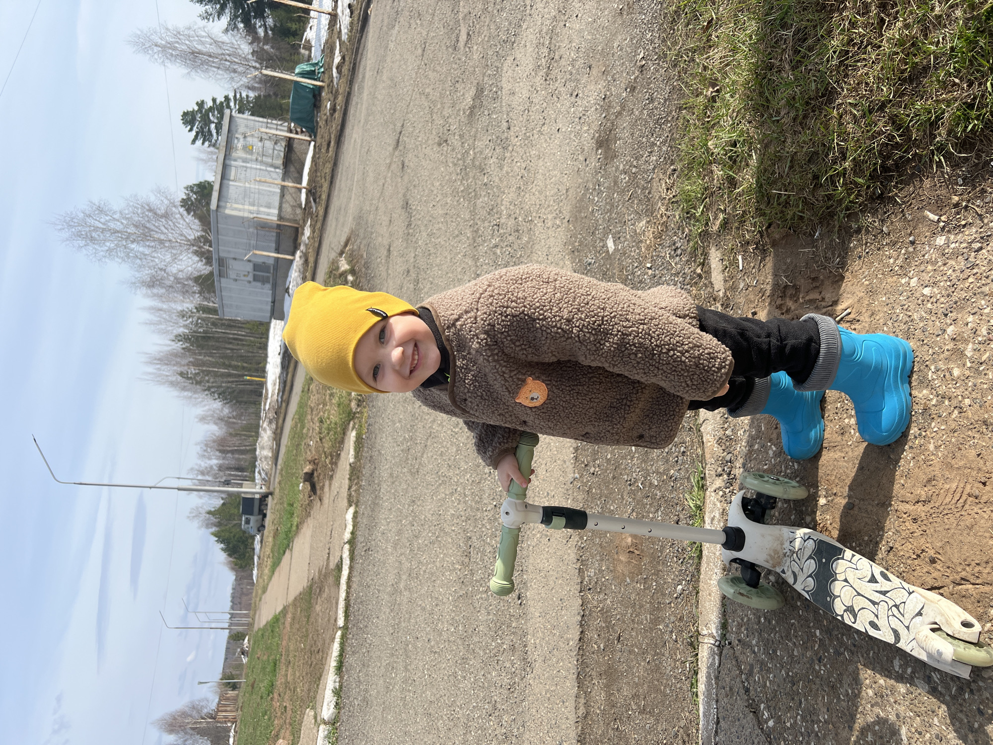 A small child stands next to a scooter on a street in Марковский, Russia. The child wears a yellow beanie, a brown teddy bear jacket with a cartoon lion patch, black pants, and blue rain boots. The scooter is white with graffiti designs on the deck. The road is asphalt with a sidewalk along the edge. A grassy area is visible to the right of the road. In the background, there are buildings, trees, and streetlights. The sky is partly cloudy.