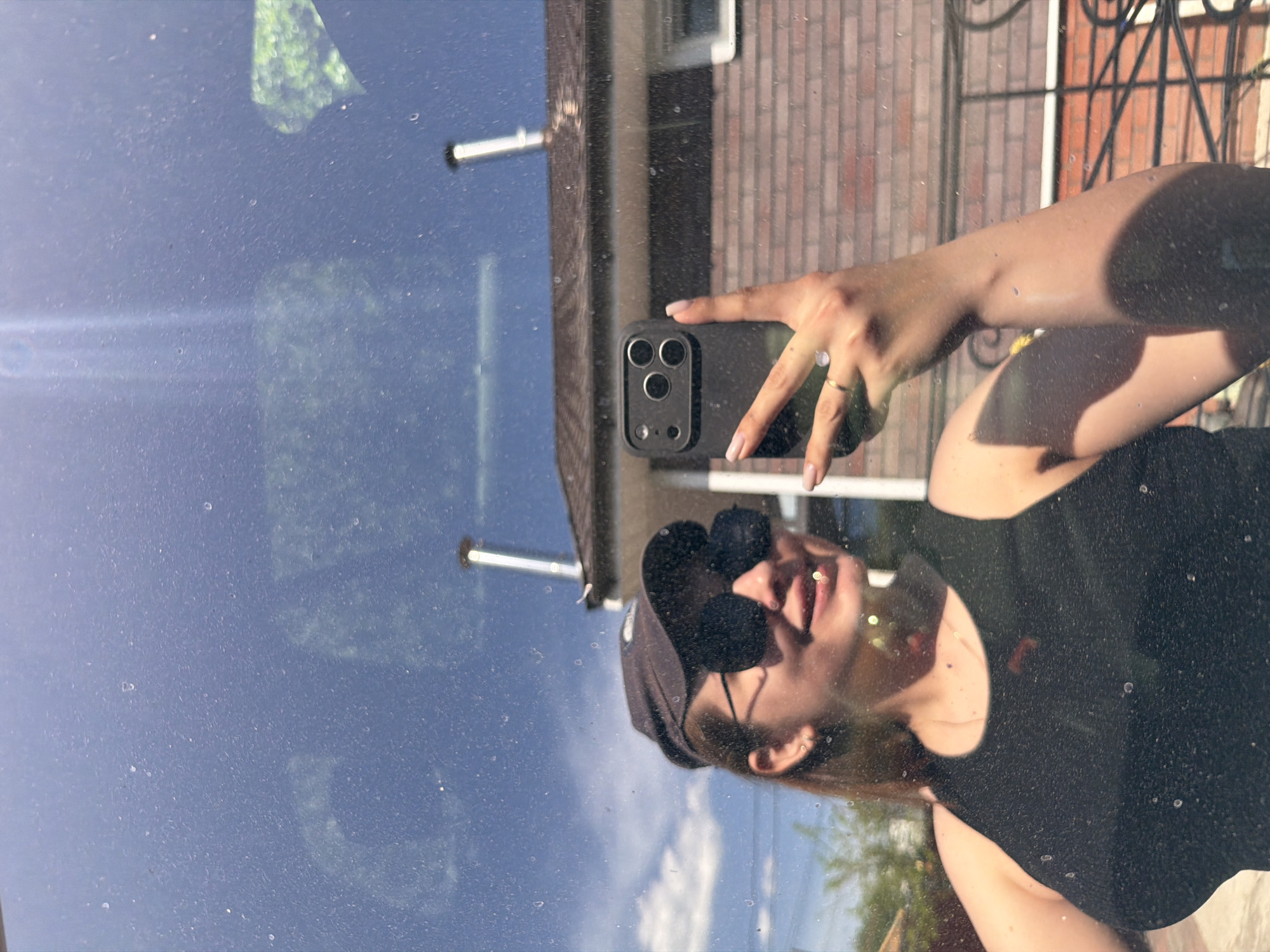 A woman is taking a selfie in a reflective surface, likely a car window, of a building in Chaikovsky, Russia. The reflection shows a brick building with a window and a section of what appears to be a metal fence or gate. The woman is wearing sunglasses and a dark-colored top, with her arm raised to hold the phone. The phone has a triple-lens camera system. The glass surface she's photographing from is speckled.