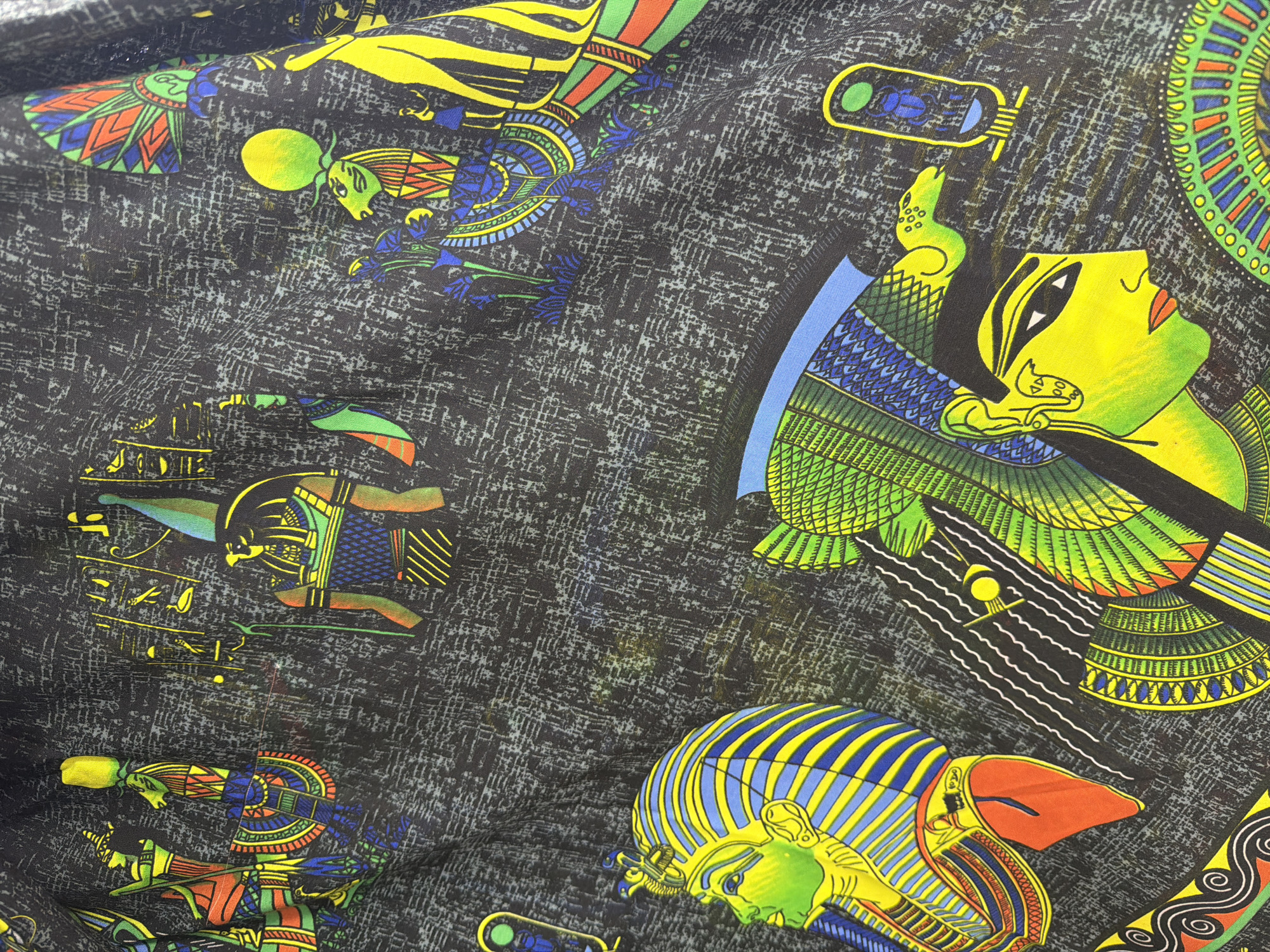 This is a close-up, top-down view of a black fabric with a vibrant, repeated pattern of Egyptian-themed imagery. The pattern includes stylized depictions of pharaohs with nemes headdresses, Egyptian gods with animal heads (such as a jackal and a falcon), hieroglyphics, and decorative borders. The colors are bold and include yellow, green, blue, red, and orange. The fabric appears to be a lightweight material, possibly cotton or polyester, and is somewhat wrinkled, indicating it might be clothing or a textile decoration. There is no discernible location in Sharm El Sheikh, Egypt, as the image solely focuses on the fabric's pattern.