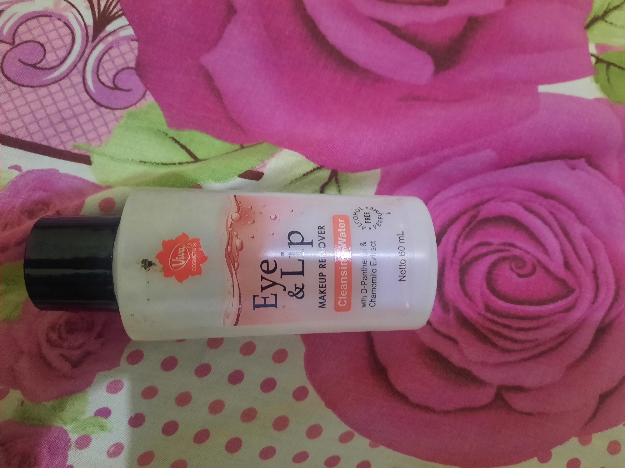 A close-up, overhead shot captures a bottle of "Viva Eye & Lip Makeup Remover Cleansing Water" resting on a patterned fabric. The fabric features a repeating pattern of pink polka dots on a light green background, interspersed with large, stylized pink roses. The bottle is white with a black cap and displays the Viva Cosmetics logo, along with the product name and ingredients. The text "Netto 60 mL" is visible at the bottom of the bottle. The lighting is natural, casting soft shadows. The image is likely taken indoors, possibly in a home environment. The city of Lhokseumawe, Indonesia, is not depicted in this image, as the scene is a close-up of a product and its background.