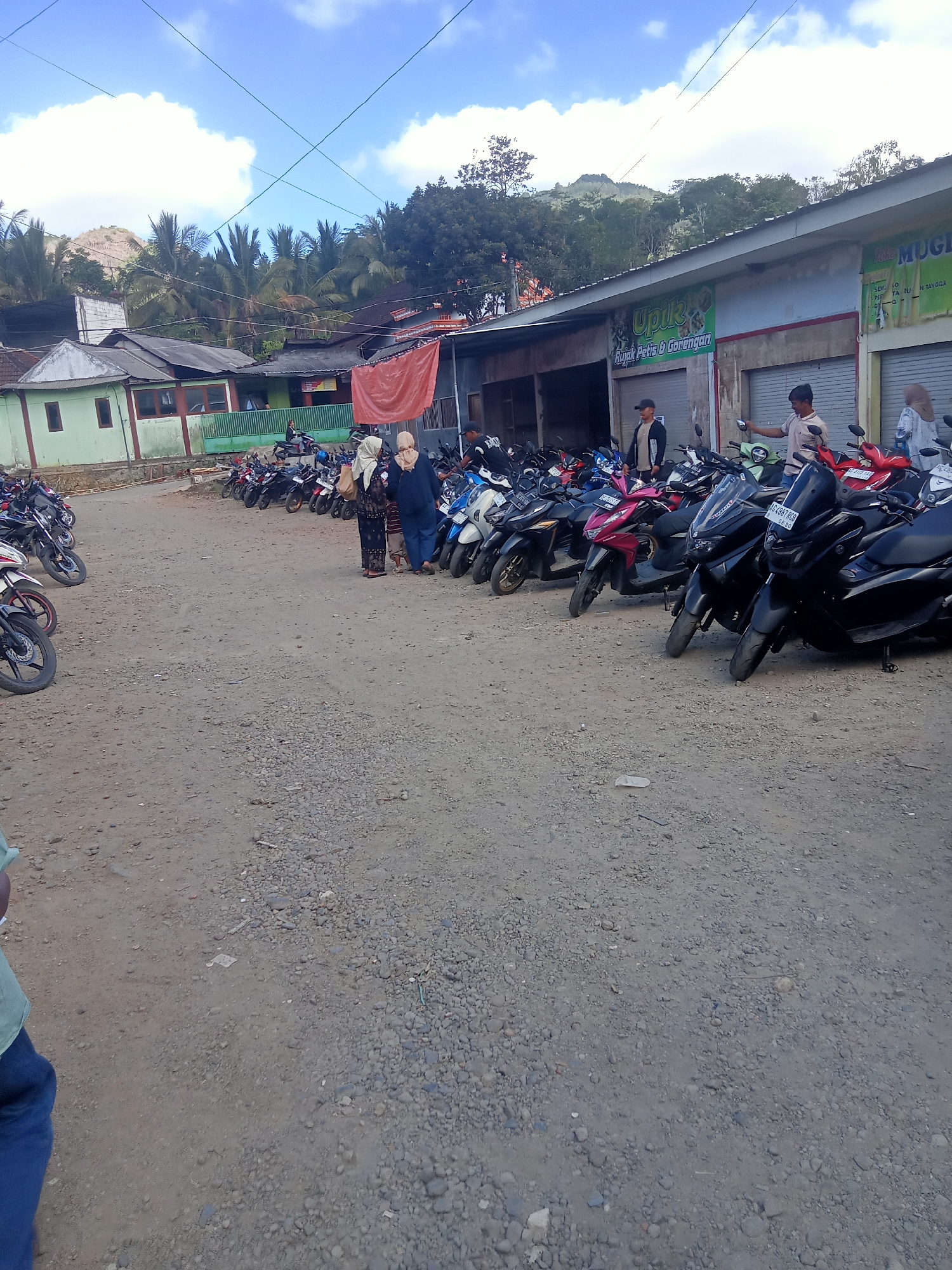 The image depicts a street scene in the city of Pule, Indonesia, with a prominent focus on a parking area filled with numerous motorcycles. The environment appears to be a mix of commercial and residential structures, with a slightly overcast sky suggesting it might be daytime.
In the foreground, the ground is a rough, gravelly dirt surface, characteristic of unpaved roads or parking lots. A significant number of motorcycles are parked in neat rows, indicating a public gathering or a busy commercial area. The motorcycles are varied in color and model, with many appearing to be scooters.
Further into the image, several buildings line the street. One building, painted light green, has visible windows and a red-tiled roof. Adjacent to it are other structures with more utilitarian facades, some featuring roller shutters, typical of shops or small businesses. Signage is visible on some of these buildings, with one sign featuring green lettering and a logo that reads "Upik" followed by "Rujak Petis & Gorengan," identifying a food establishment. Another sign, partially visible, reads "MUGI."
There are a few individuals present in the scene. Two women, both wearing headscarves, are standing near the parked motorcycles in the middle ground, appearing to be conversing. One woman is dressed in a dark patterned outfit, while the other wears a lighter colored headscarf and a dark top. To their right, a man in a light-colored t-shirt and dark pants is interacting with a motorcycle. In the background, a man wearing a cap and a jacket is standing near some of the parked motorcycles. Another man in a white t-shirt is visible further back, standing near the entrance of a building.
The background shows a lush, hilly landscape with numerous trees, including palm trees, suggesting a tropical or sub-tropical climate. Overhead, wires crisscross the sky, common in urban and semi-urban settings. The overall impression is a snapshot of everyday life in a community in Pule, Indonesia, where motorcycles are a primary mode of transportation and commercial activity is ongoing.