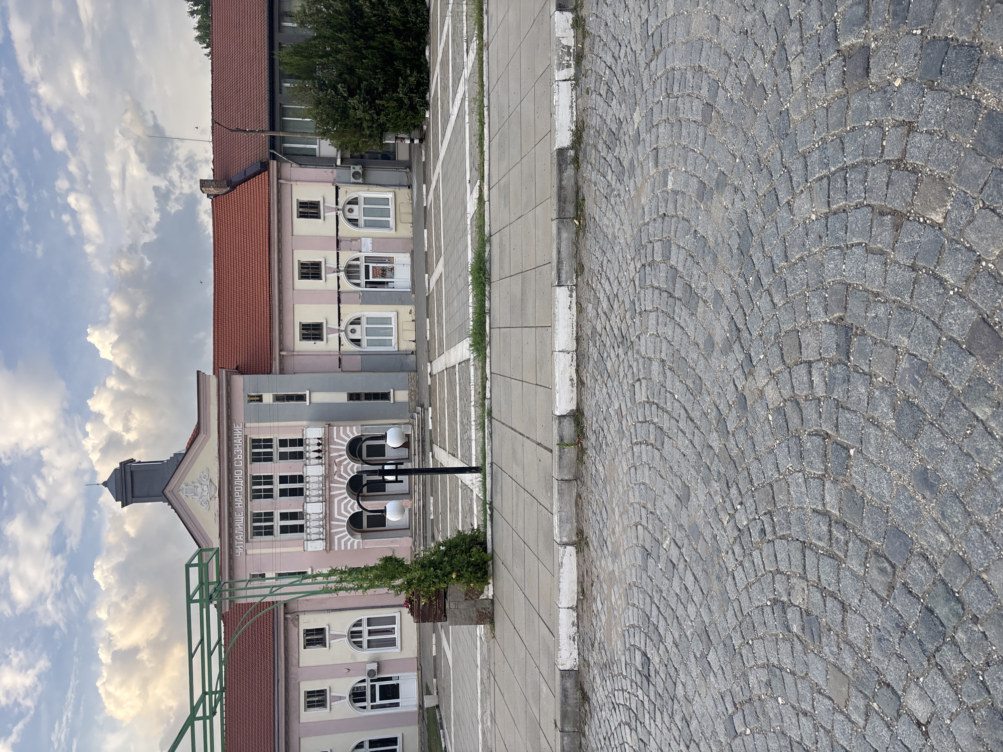 The image depicts the exterior of the Community Cultural Center "Narodno Saznanie" in the city of Breznitsa, Bulgaria. The building features a light pink facade with arched windows and entrances. A balcony with ornate white railings is visible on the upper floor. Above the entrance, text in Cyrillic reads "ЧИТАЛИЩЕ. НАРОДНО СЪЗНАНИЕ" (Community Cultural Center "Narodno Saznanie"). A clock tower with a dark gray roof and spire rises from the center of the building. To the right of the main building, a gray structure with a dark red tiled roof is attached. Green metal archways and a tall, slender evergreen tree are present in front of the building. The foreground is paved with cobblestones, and a sidewalk made of concrete tiles with white-bordered edges separates it from a paved area in front of the cultural center. The sky is partly cloudy, with soft, diffused sunlight illuminating the scene. No people are visible in the image.