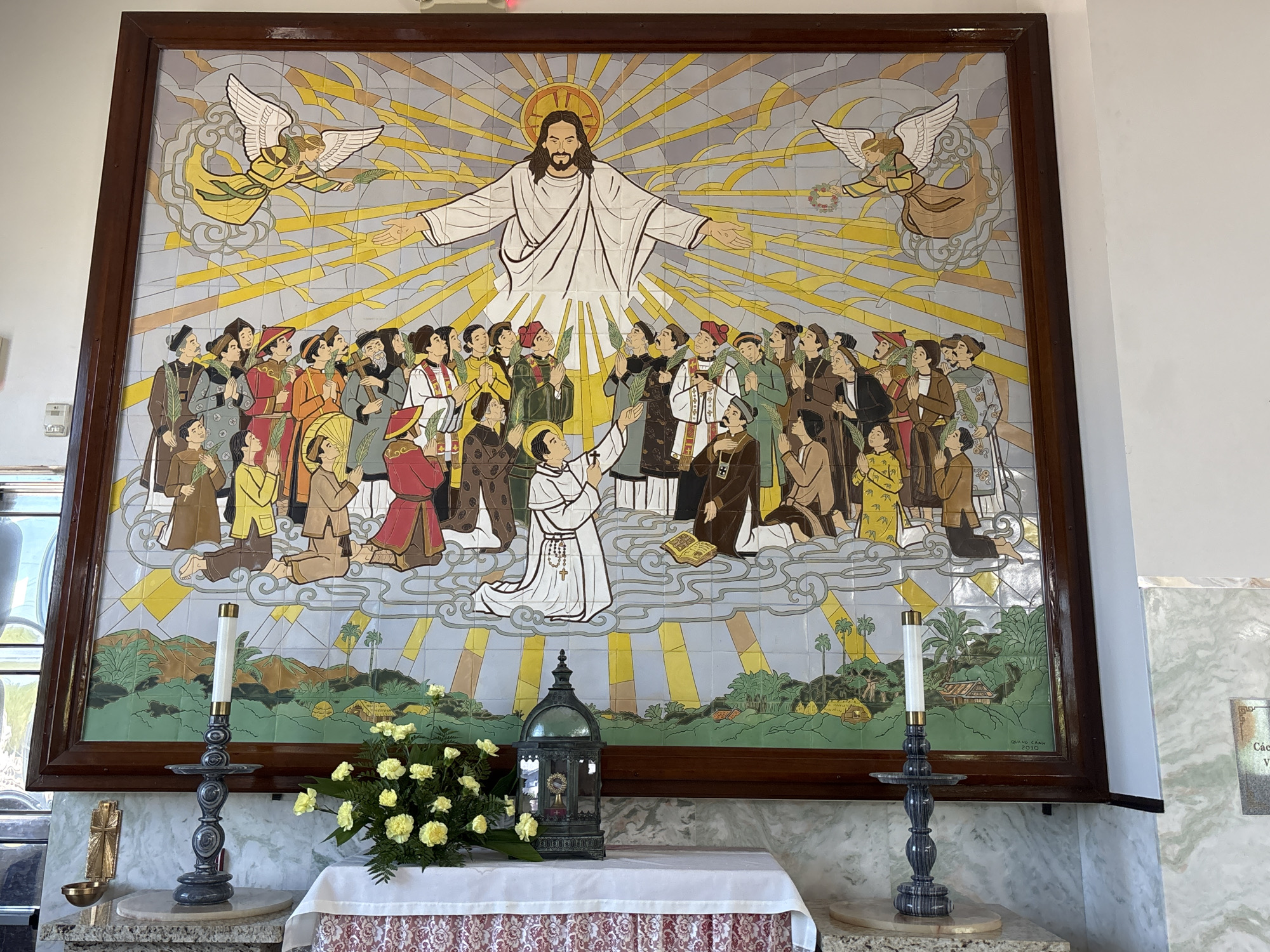 This image depicts a religious mural in Phoenix, Arizona, United States of America, located within a building that appears to be a church or chapel. The mural, framed by a dark wood border, is a large tile mosaic art piece. At the center of the mural, a figure resembling Jesus Christ stands with arms outstretched, radiating golden light from a halo. He is depicted wearing a white robe. Flanking Jesus are two angels with outstretched wings, one on the left holding a palm frond and the other on the right holding a wreath. Below Jesus, a crowd of people in various attire, some wearing traditional Vietnamese conical hats, are gathered. Many in the crowd are holding palm fronds and have their hands clasped in prayer. A central figure in a white robe with a rosary is kneeling and holding a book. The background of the mural features stylized clouds and a landscape with greenery and small houses.
In the foreground, below the mural, is an altar area. A white tablecloth with a red and white patterned trim covers the altar. On the altar, there are several objects: a decorative dark metal lantern containing a monstrance, a bouquet of yellow flowers, and two candle holders with lit white candles. To the left of the altar, on a marble surface, is a gold-colored cross and a small bowl. The walls in the background are a light color, and a section of marble wall with green veining is visible to the right. The overall impression is one of a sacred space, likely for worship or remembrance.