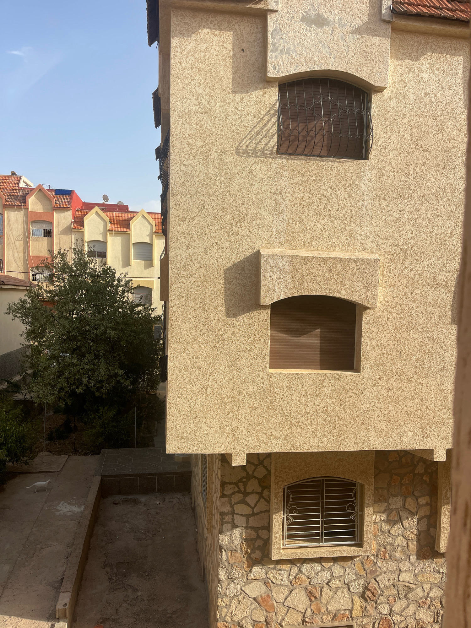 This image captures a segment of a multi-story building in the city of Azrou, Morocco, under a clear sky. The building features a rough, textured beige exterior on its upper floors, with arched window openings. One window on the upper floor is covered by a metal grate with decorative scrollwork, and a brown roller blind. Another window on a slightly lower level also has a brown roller blind, and a stone architectural element above it. The lower portion of the building is constructed with rough-hewn stones of varying sizes and colors, and features a barred window with a white frame. Adjacent to the building on the left is another structure with a red tiled roof and a similar architectural style, partially obscured by a leafy green tree. In the foreground, a paved area with some dirt and a low concrete border is visible. A white cat is seen walking on a paved pathway to the left. The overall scene suggests a residential or commercial area in a Moroccan city, with typical architectural features and a domestic animal present.