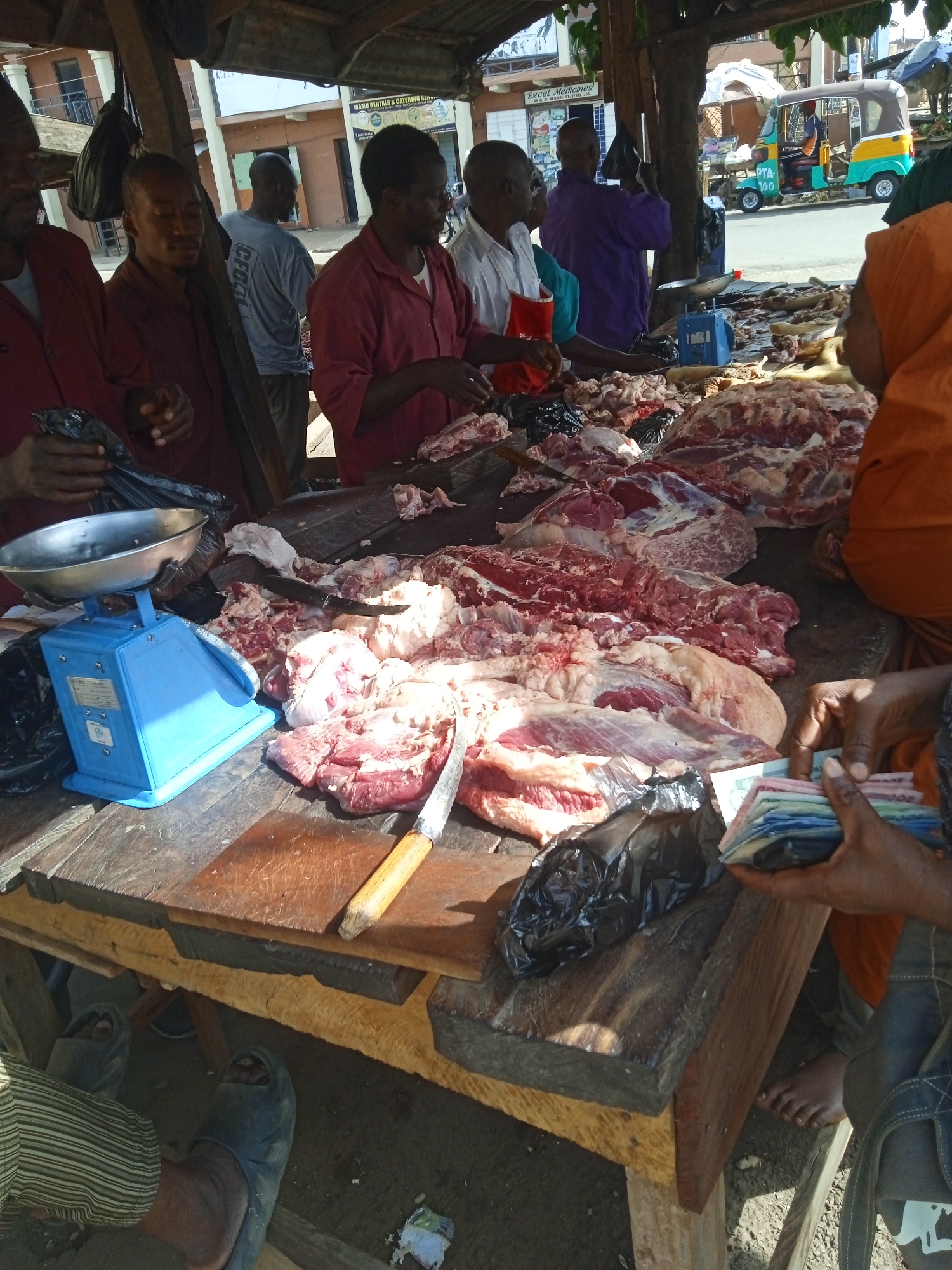 The image depicts an outdoor butcher shop scene in Jos, Nigeria. Several men stand behind a wooden table covered with large cuts of raw meat. A blue weighing scale is on the table along with a knife. A person in an orange garment holds a stack of banknotes. Other people are visible around the table. A road and buildings are in the background, including a three-wheeled vehicle. The scene is under a corrugated metal roof supported by wooden posts.