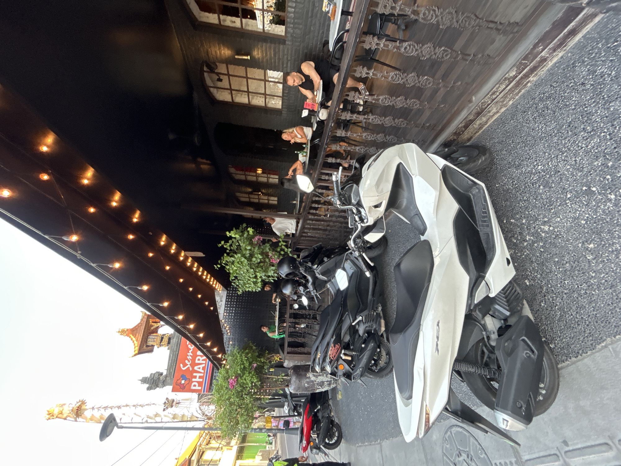 In North Kuta, Indonesia, this image captures a street scene outside a restaurant with an outdoor seating area. Several scooters are parked on the sidewalk. A white scooter is prominently in the foreground, facing left. Behind it, a black scooter is parked, and further back, a red scooter is visible. The restaurant features a dark facade with large windows and an overhang adorned with decorative lights. People are seated at tables on the restaurant's patio, some engaged in conversation or eating. The background shows a street with buildings and a sign that reads "Seminyak Pharma." The lighting suggests it is late afternoon or early evening.