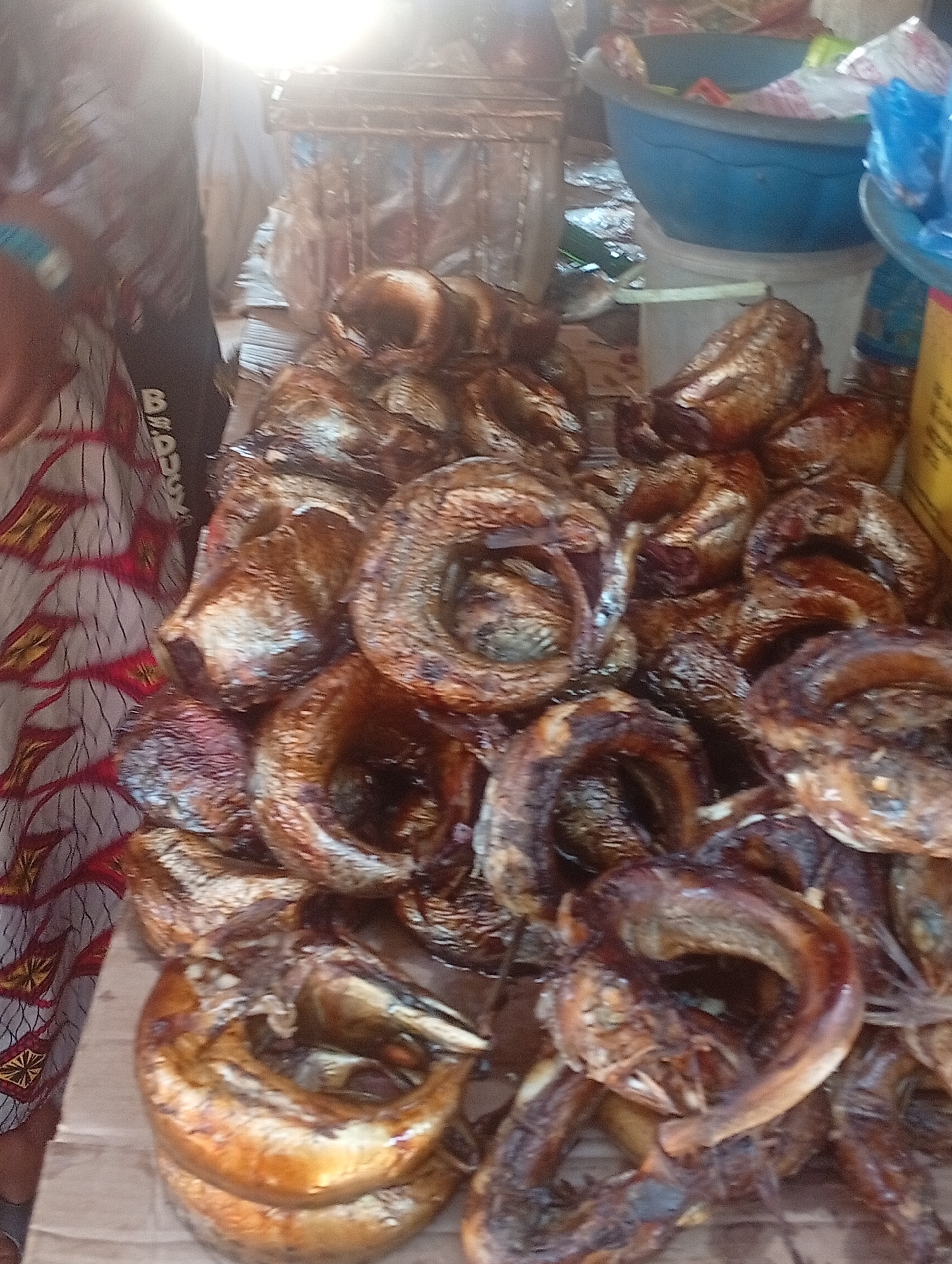 The image depicts a market stall piled high with smoked fish. The fish appear to be catfish, identifiable by their elongated bodies and lack of scales. They are arranged on a piece of cardboard on a table, likely for sale. The stall is set up outdoors in an unknown area of a city. A person wearing a patterned fabric is partially visible to the left. A blue container sits in the background, possibly used for carrying or displaying other goods. The setting is a typical open-air market.