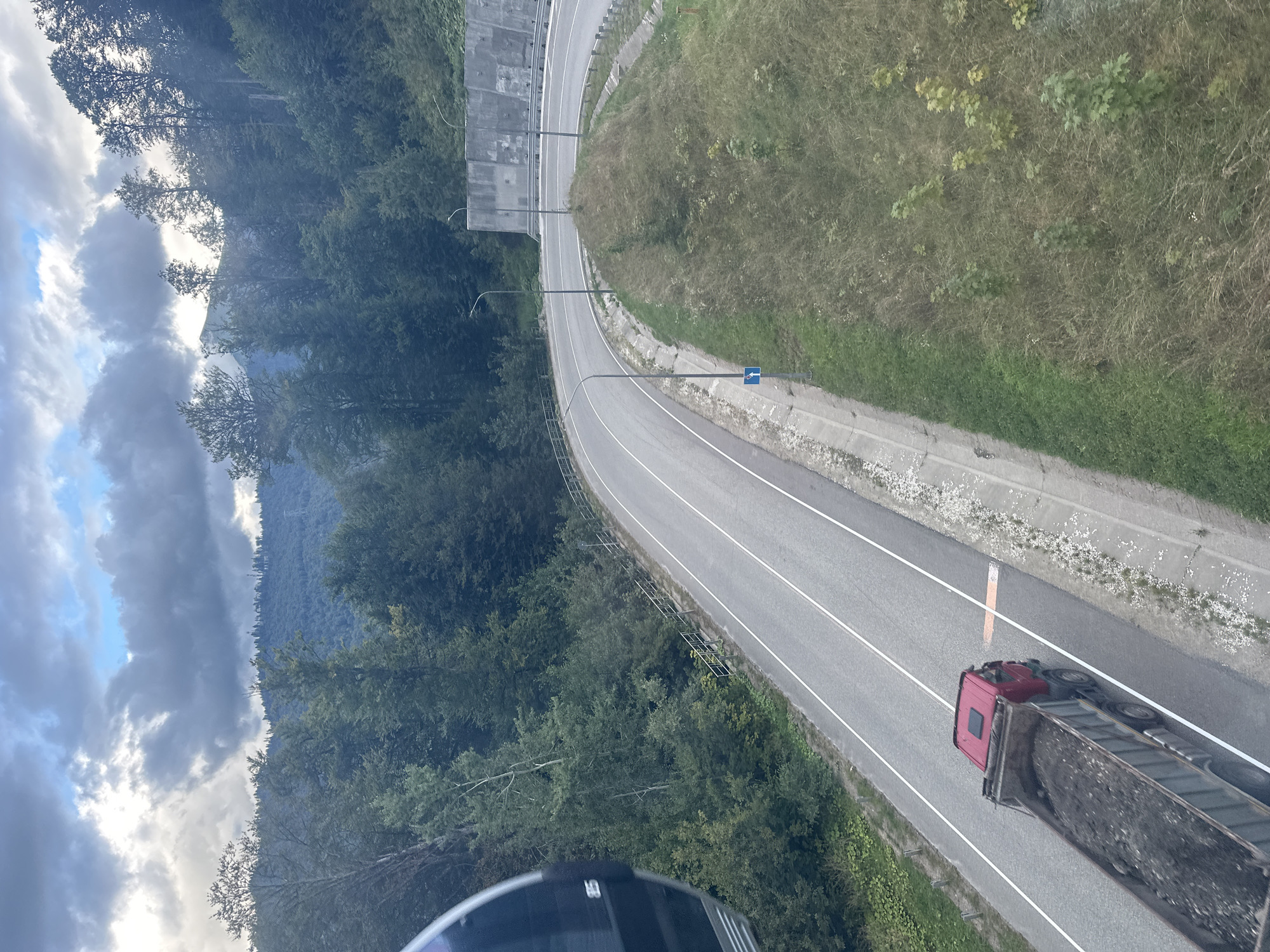 The image depicts a scenic view from a cable car in Estosadok, Russia, looking down at a winding asphalt road through a mountainous, forested area. A red truck loaded with dark soil is traveling on the road. The road is flanked by green vegetation and a concrete retaining wall on the right side, with a metal guardrail. The sky is partially cloudy with hints of blue. The cable car cabin is partially visible in the upper left corner of the frame.